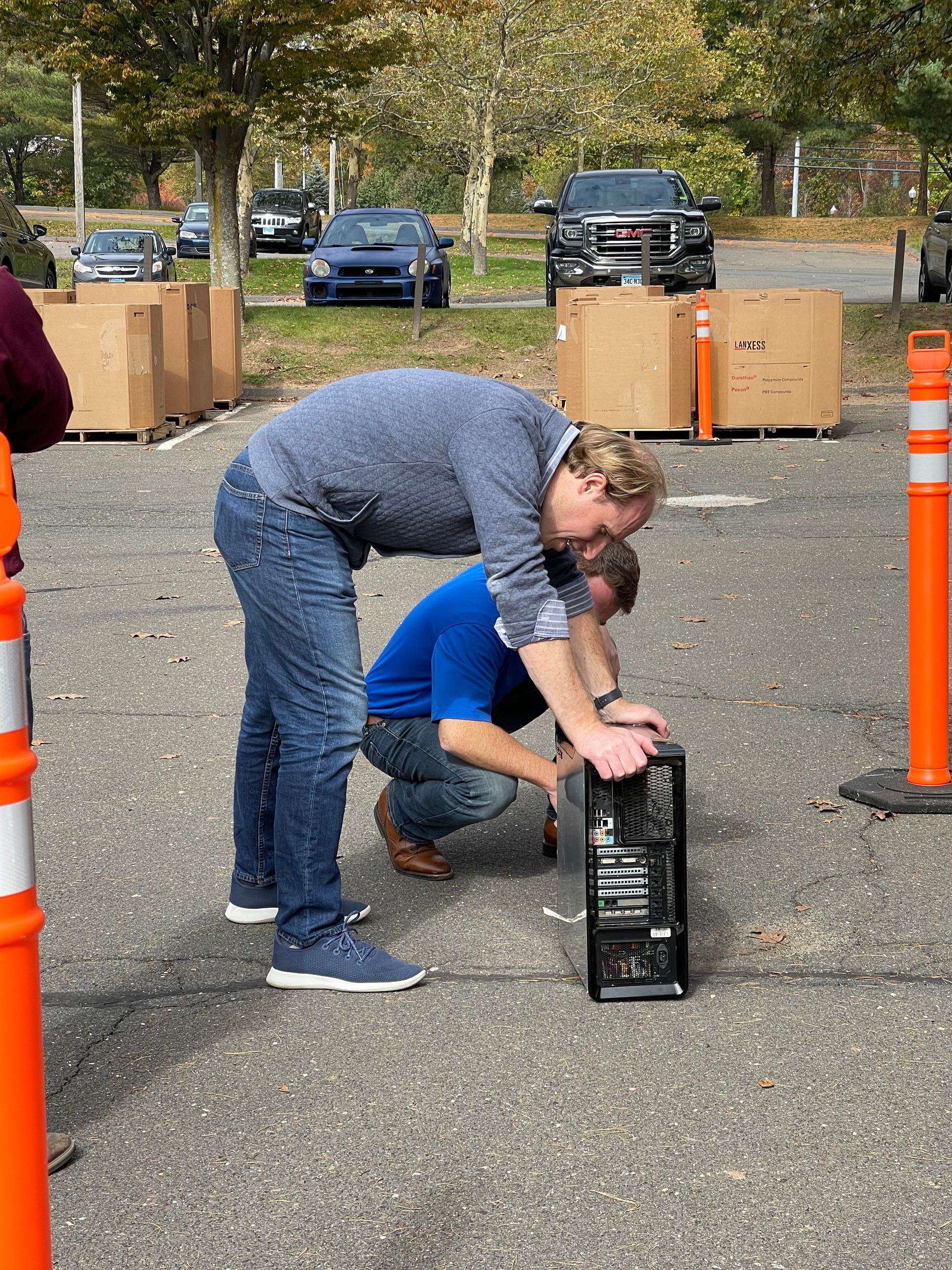 Two people crouch beside a black crate on pavement, with orange cones and boxes in the background.