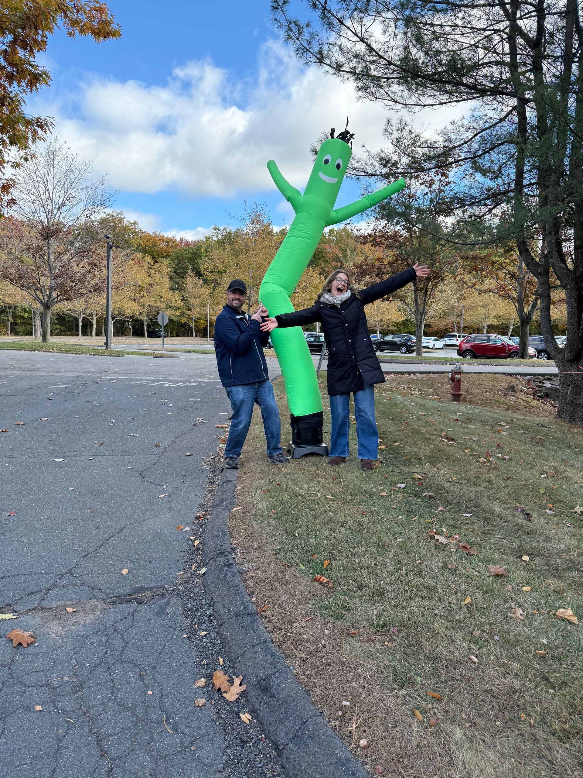 Two people pose beside a green inflatable tube man in a parking lot with autumn trees and blue sky.