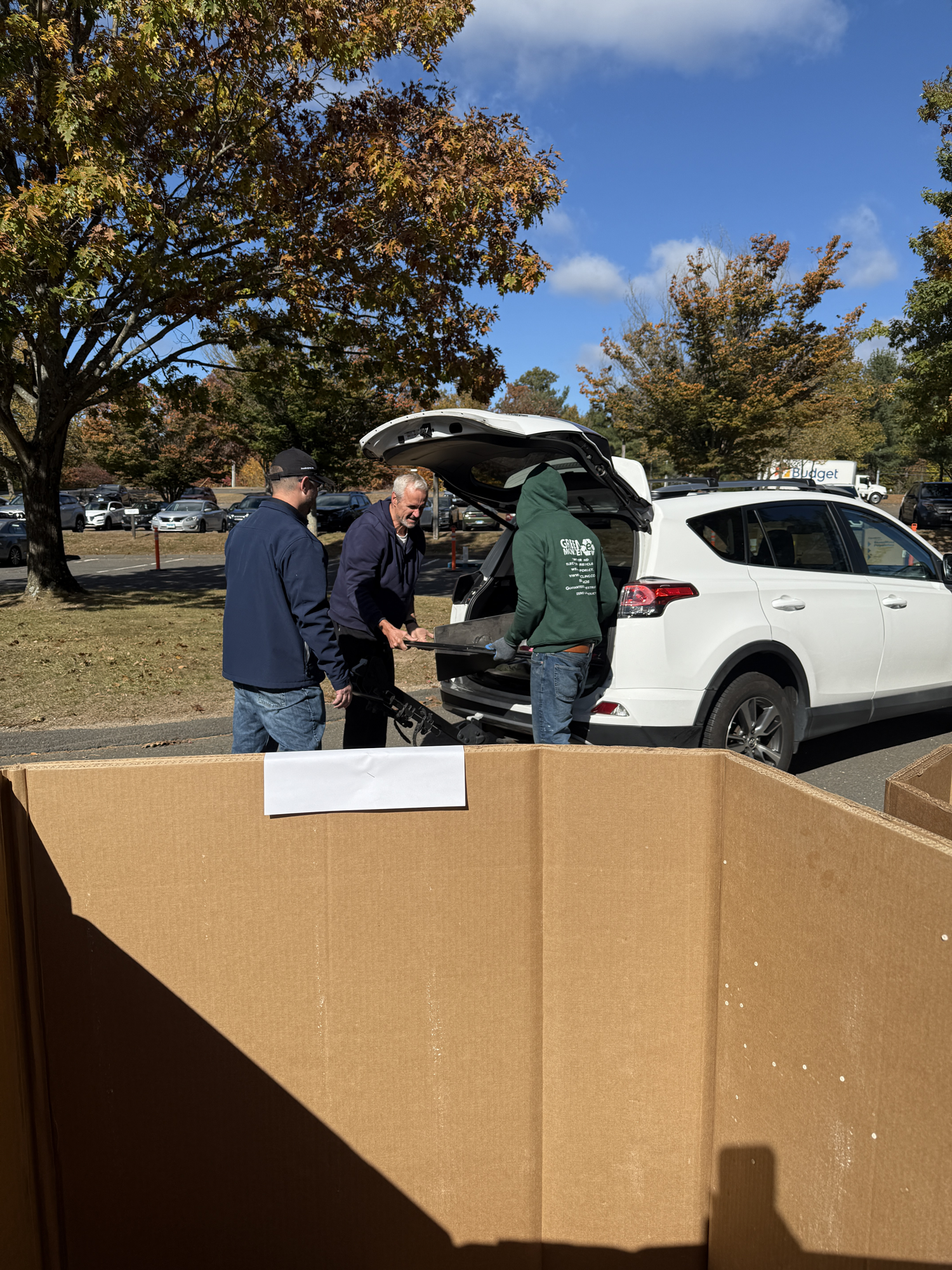 People unloading items from SUVs into a large cardboard bin outdoors on a sunny day.