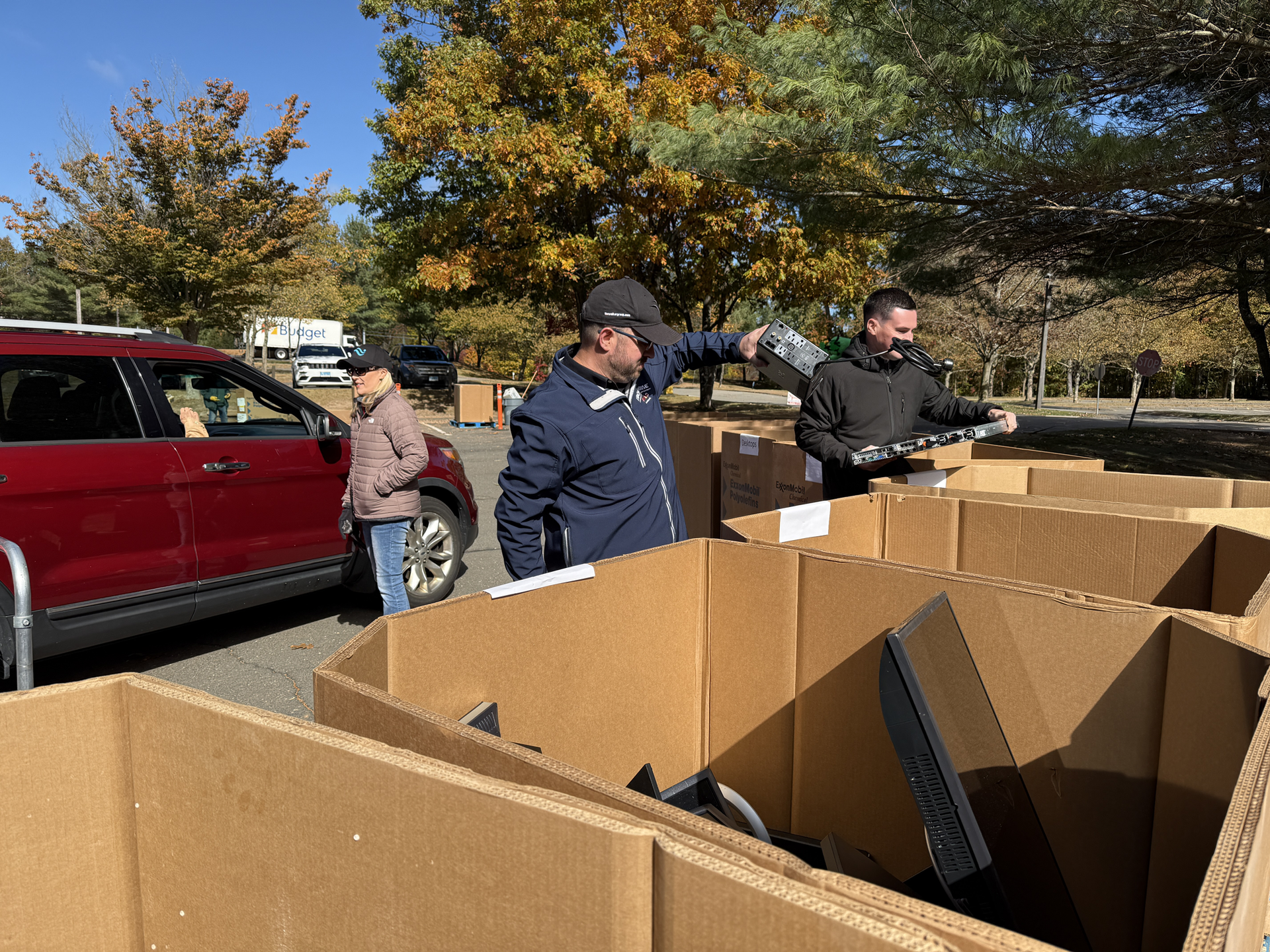 People sorting cardboard boxes outdoors beside a red SUV in a sunny parking lot.