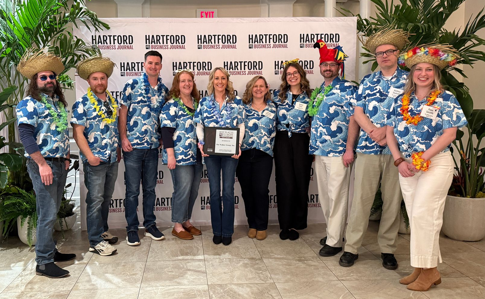 Group in blue Hawaiian shirts posing indoors in front of a sign, with a plant on the left and festive hats.
