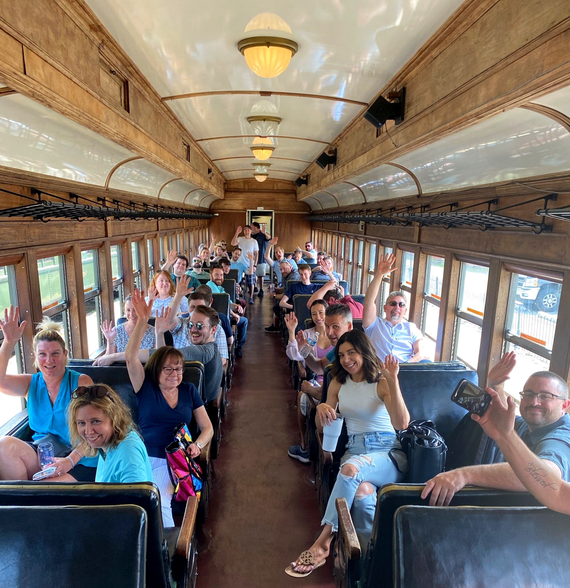 Passengers waving inside a vintage train car with wooden seats and arched ceiling.