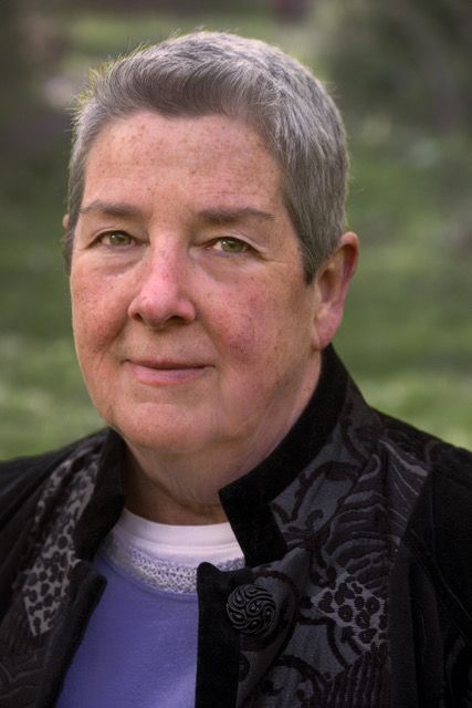 Person with short gray hair wearing a dark jacket, looking at the camera outdoors with a blurred green background