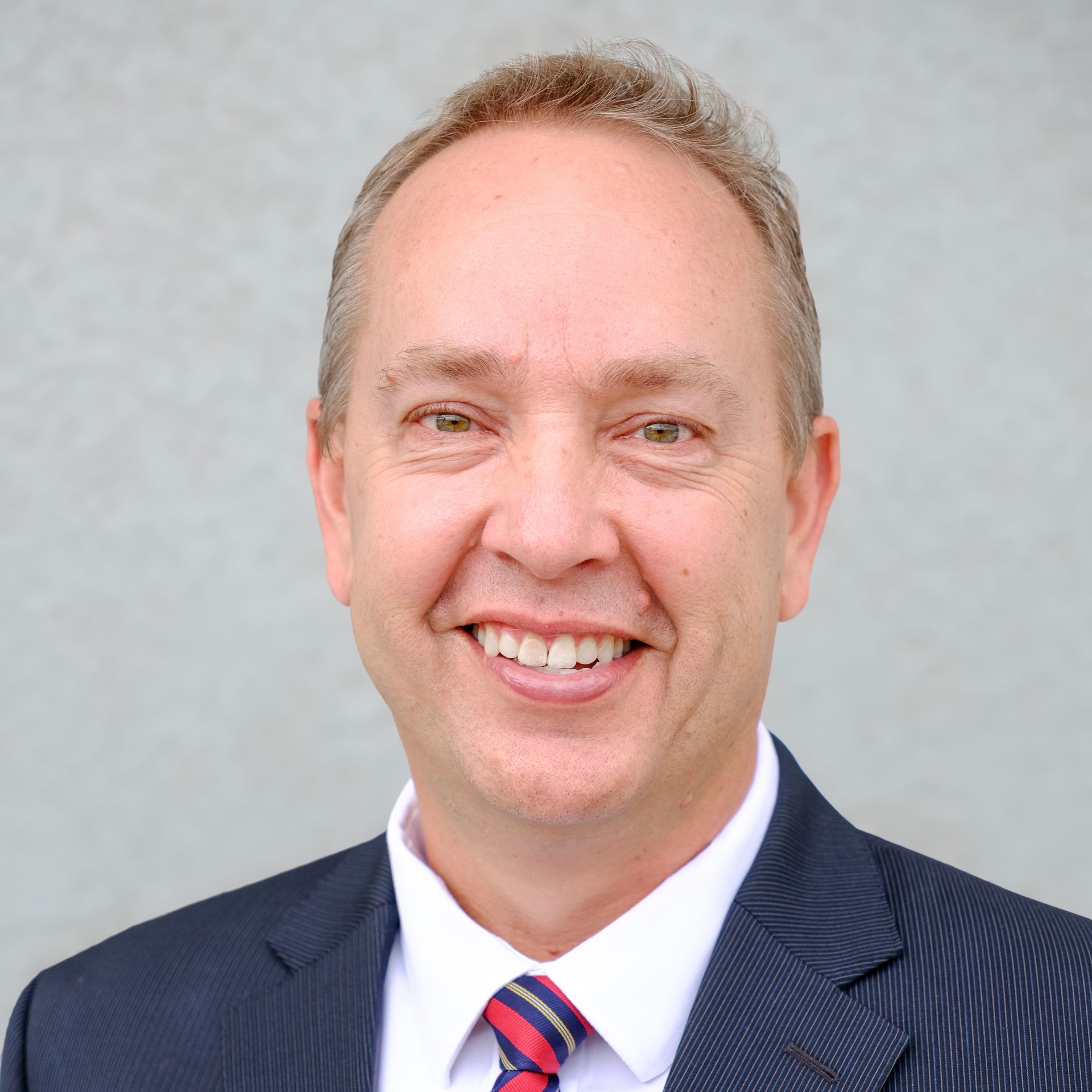 Portrait of a smiling man in a navy suit and striped tie against a light gray background