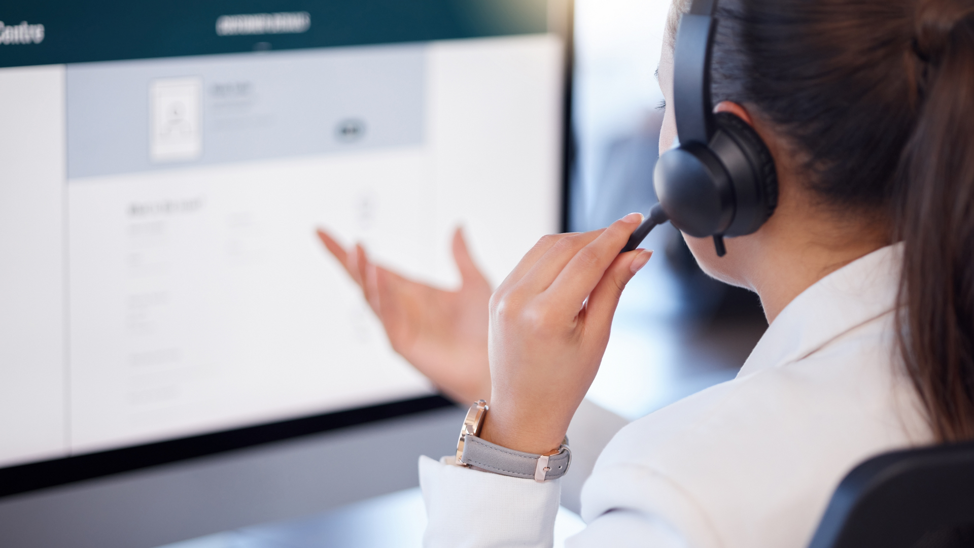 Call center agent wearing a headset, gesturing toward a computer screen in an office.