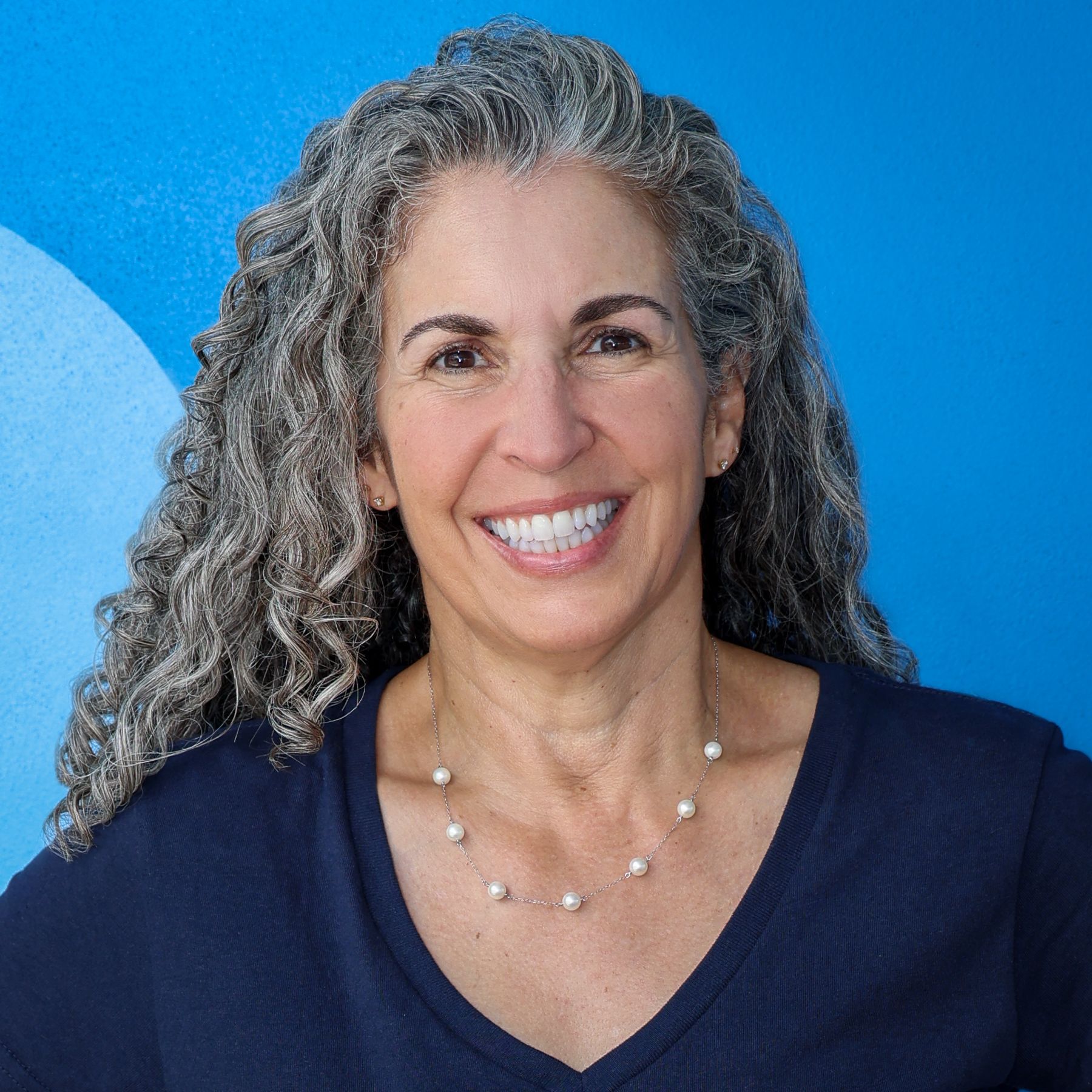 Smiling woman with curly gray hair wearing a navy top against a bright blue background
