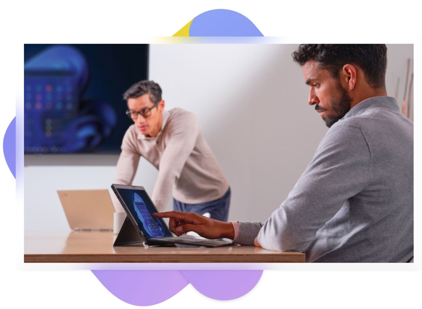 Two people working at a laptop in a meeting room, one seated and one leaning in to talk