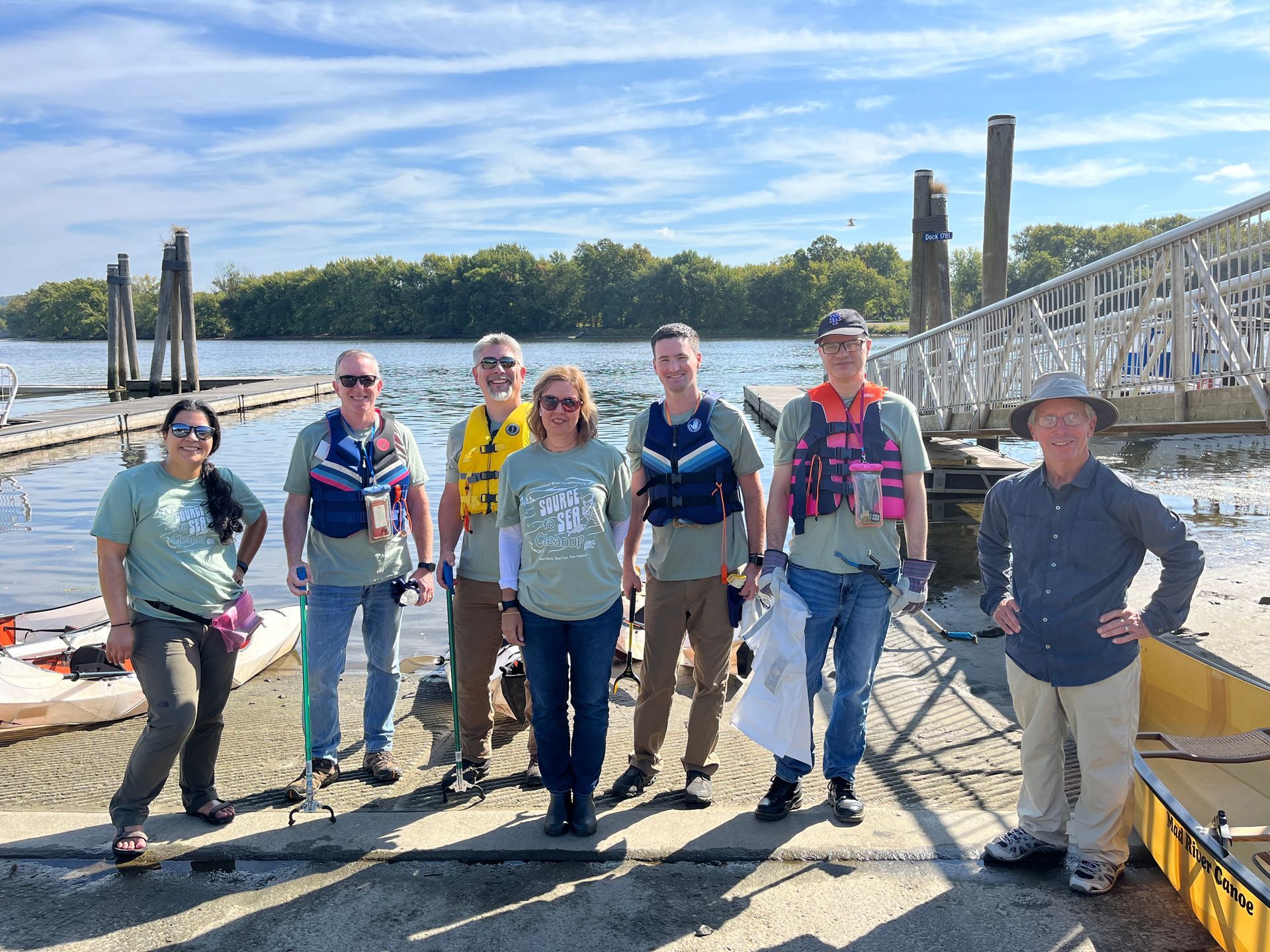 Seven people standing on a dock with boats, near a white bridge on a sunny day.