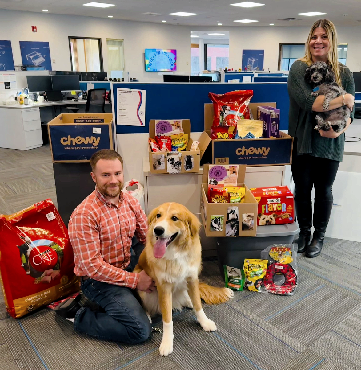 Two people and a dog pose with Chewy pet donations in an office.