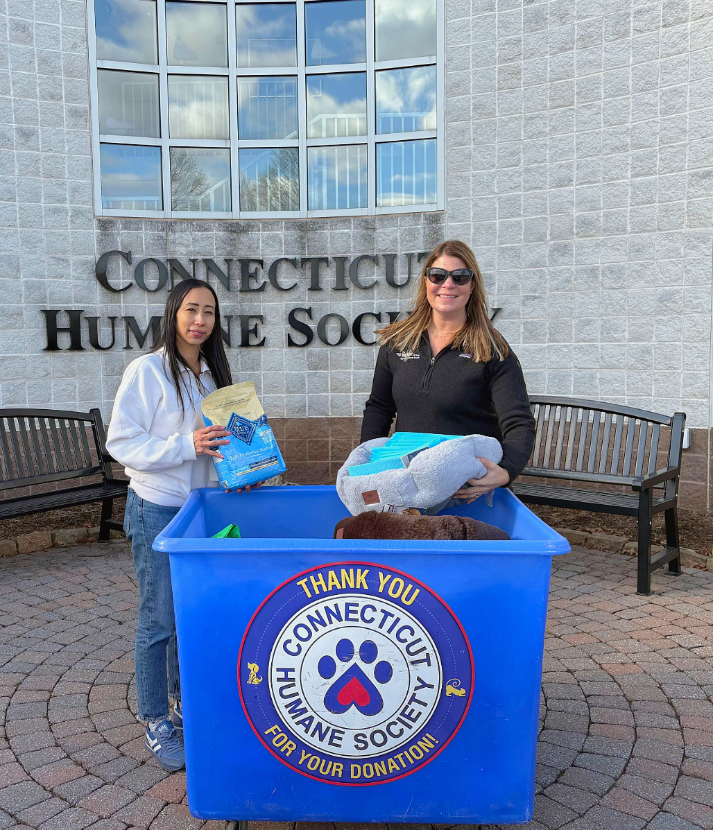 Two women stand by a blue donation bin outside Connecticut Humane Society, holding pet supplies.