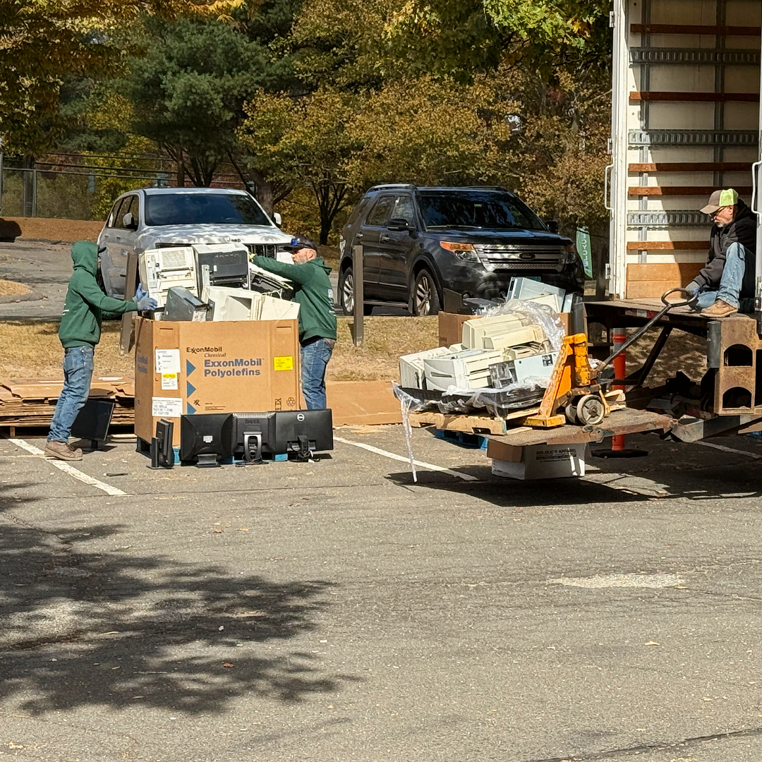 Workers loading equipment onto a trailer in an outdoor lot, with parked trucks and autumn trees nearby