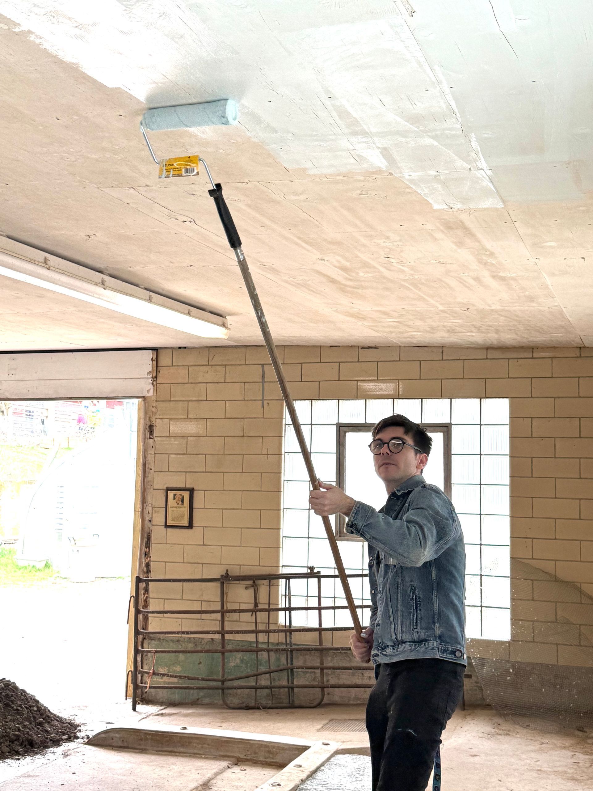 Worker painting a ceiling with a roller in an unfinished room, standing near exposed brick walls.