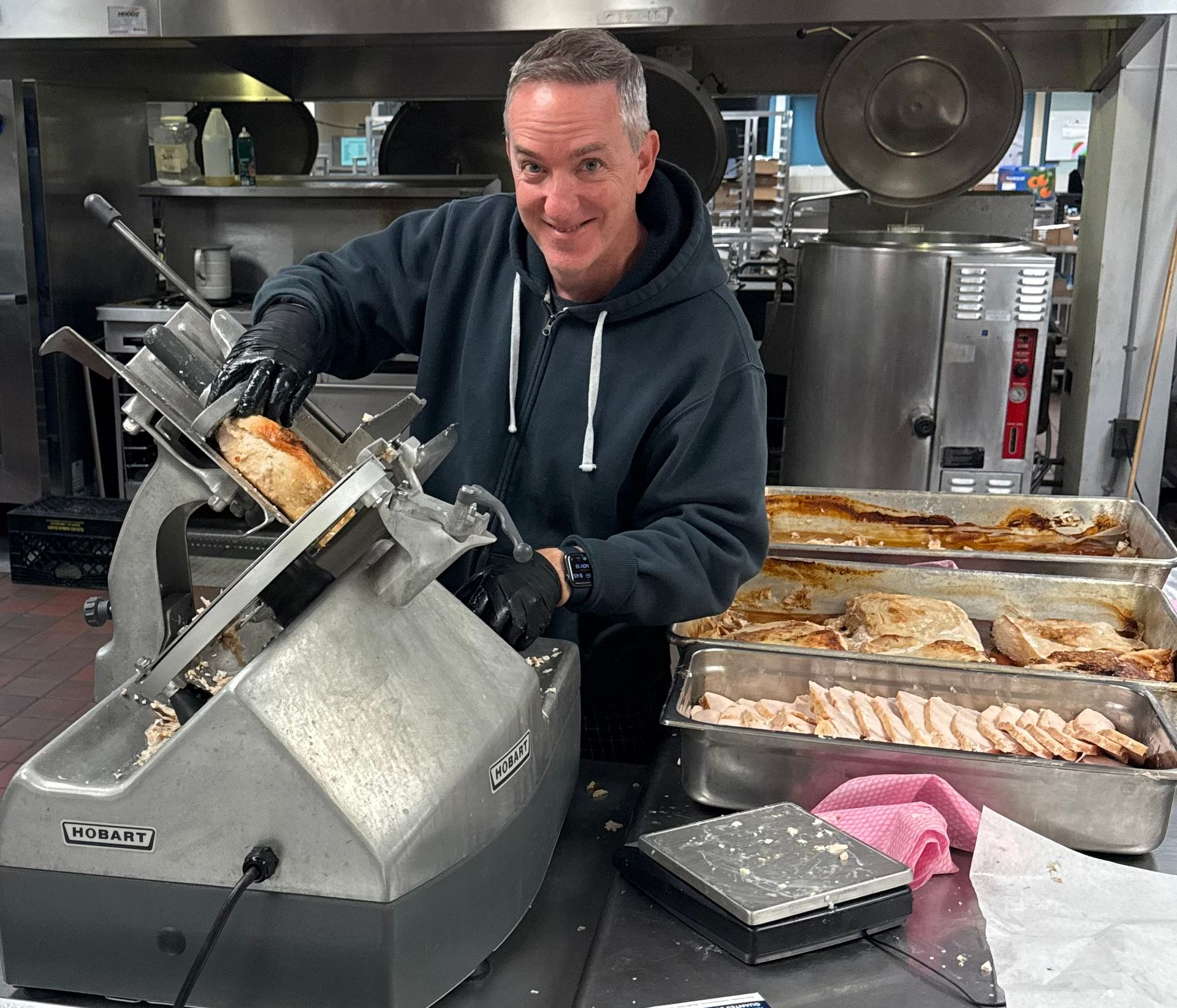 Man slicing meat in a commercial kitchen beside trays of prepared food