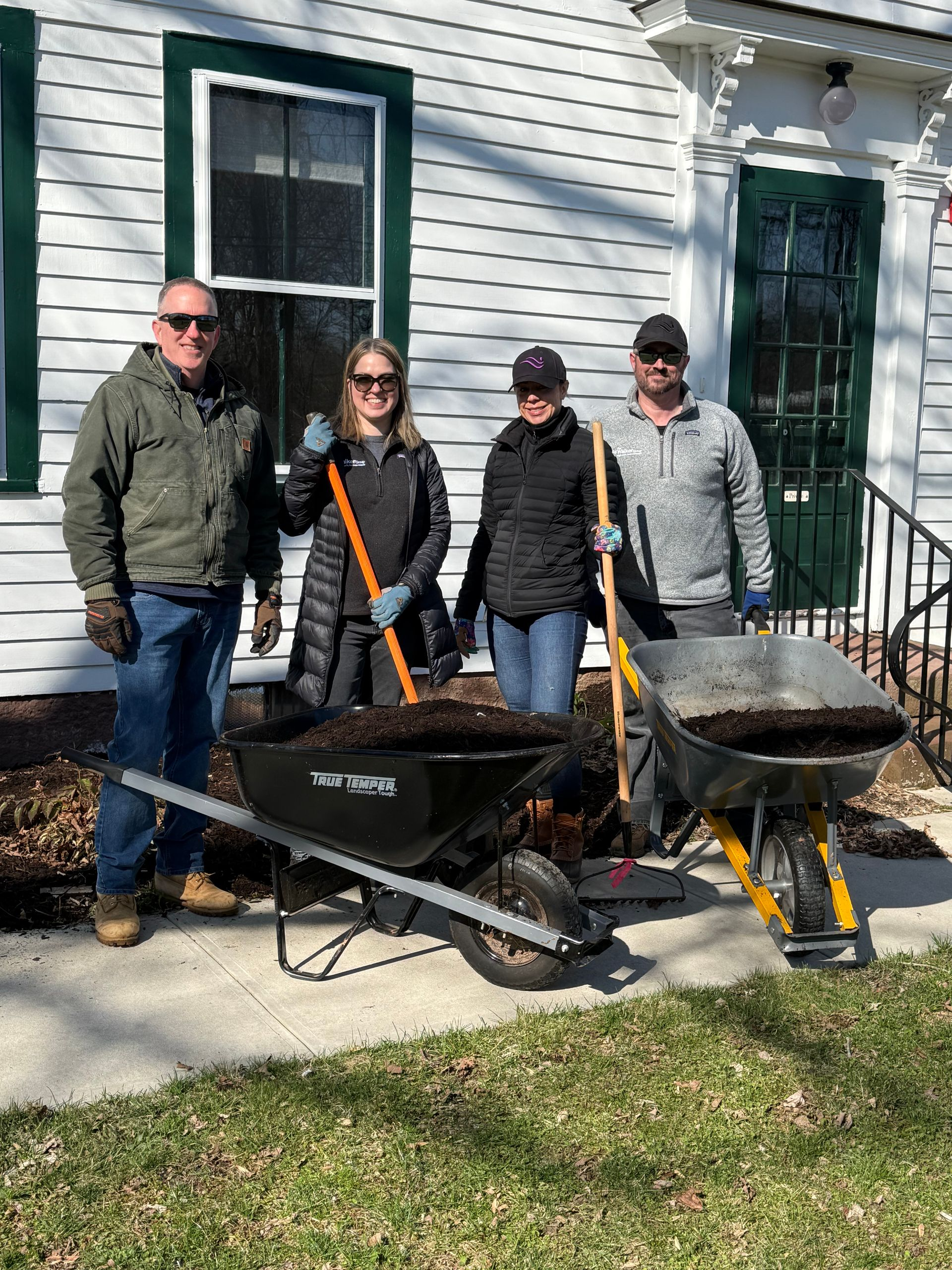 Four people standing behind two wheelbarrows full of dirt outside a white house