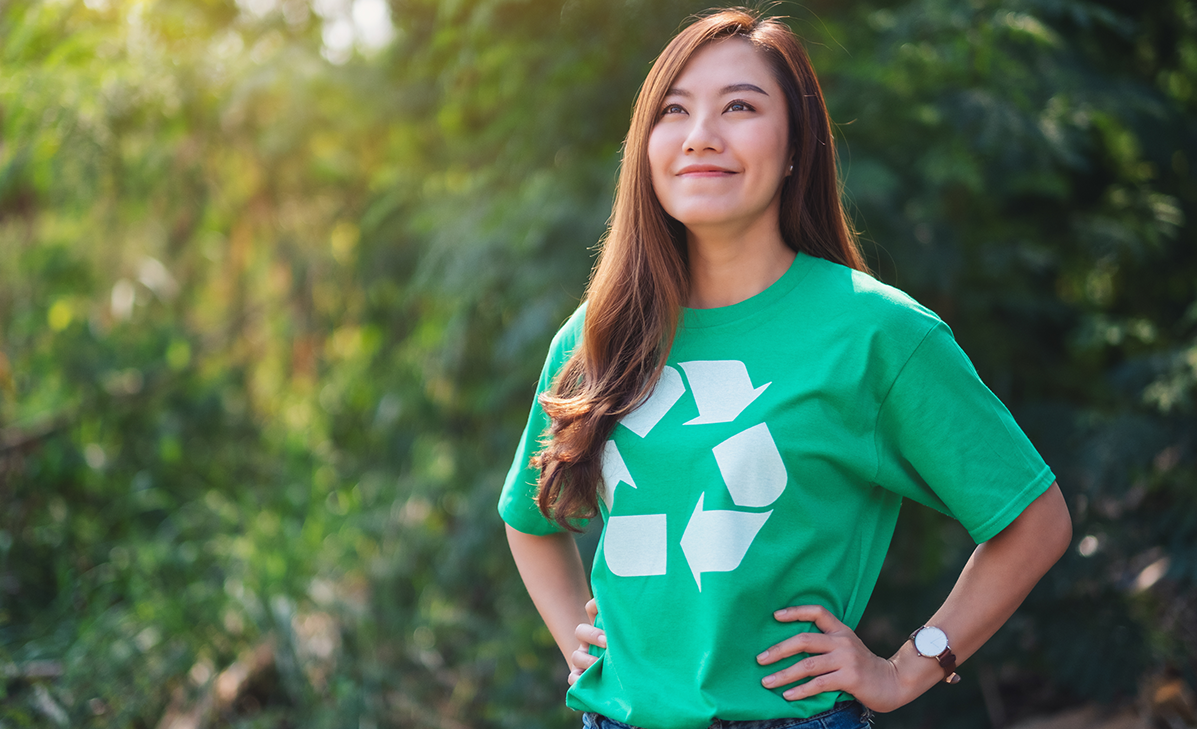 Woman in a green T-shirt standing outdoors with hands on hips, smiling in a sunlit park.