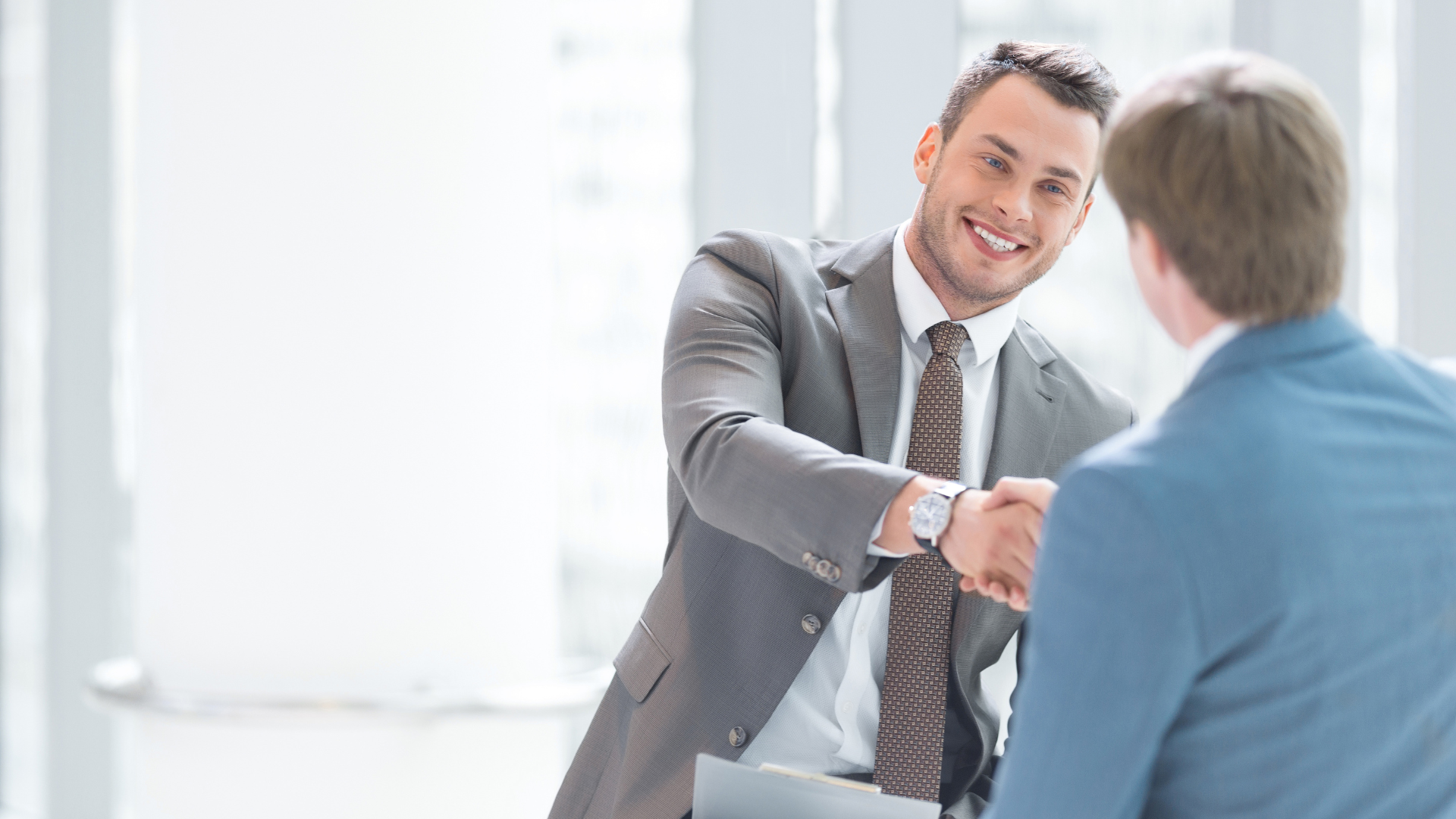 Man in gray suit shaking hands with another person, both smiling in a bright office.