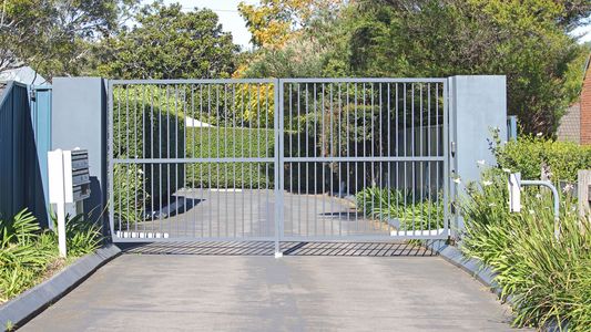 A metal gate is open to a driveway with trees in the background.