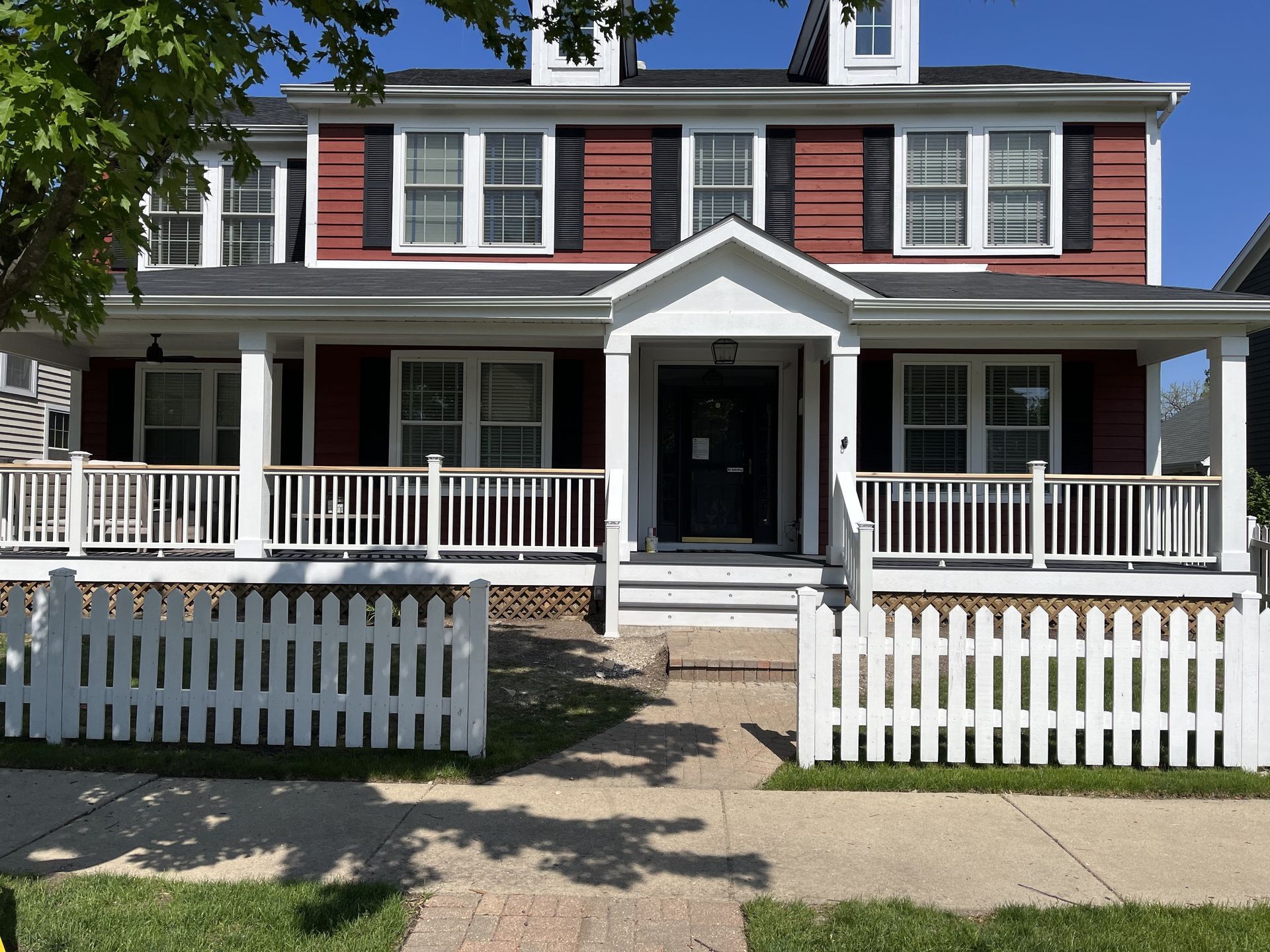 Two-story red house with white porch, black shutters, and a white picket fence
