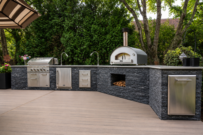Outdoor kitchen with stone counter, stainless grill, pizza oven, and sink on a patio
