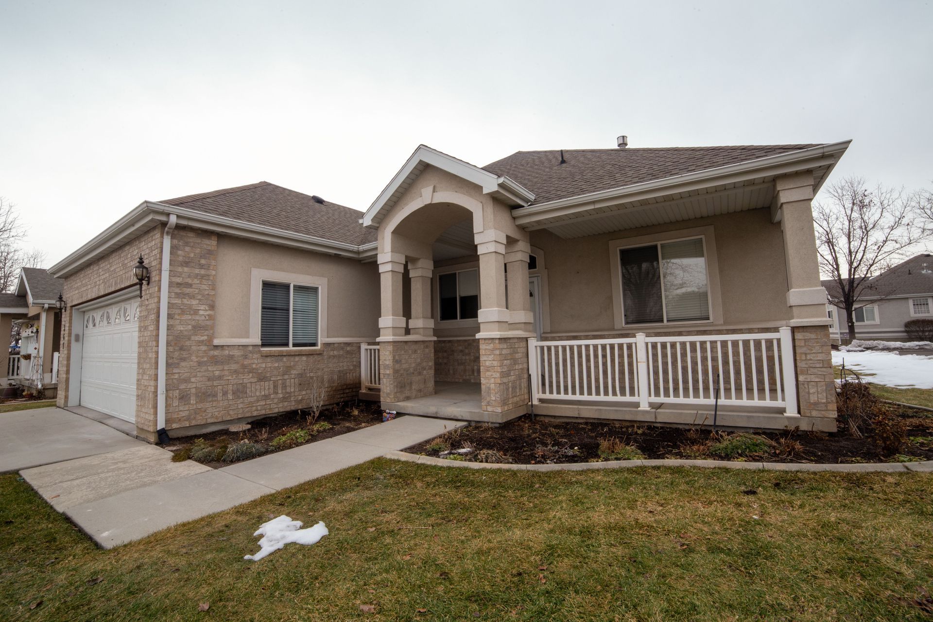 Single-story beige house with a front porch, white railing, and a driveway on an overcast day