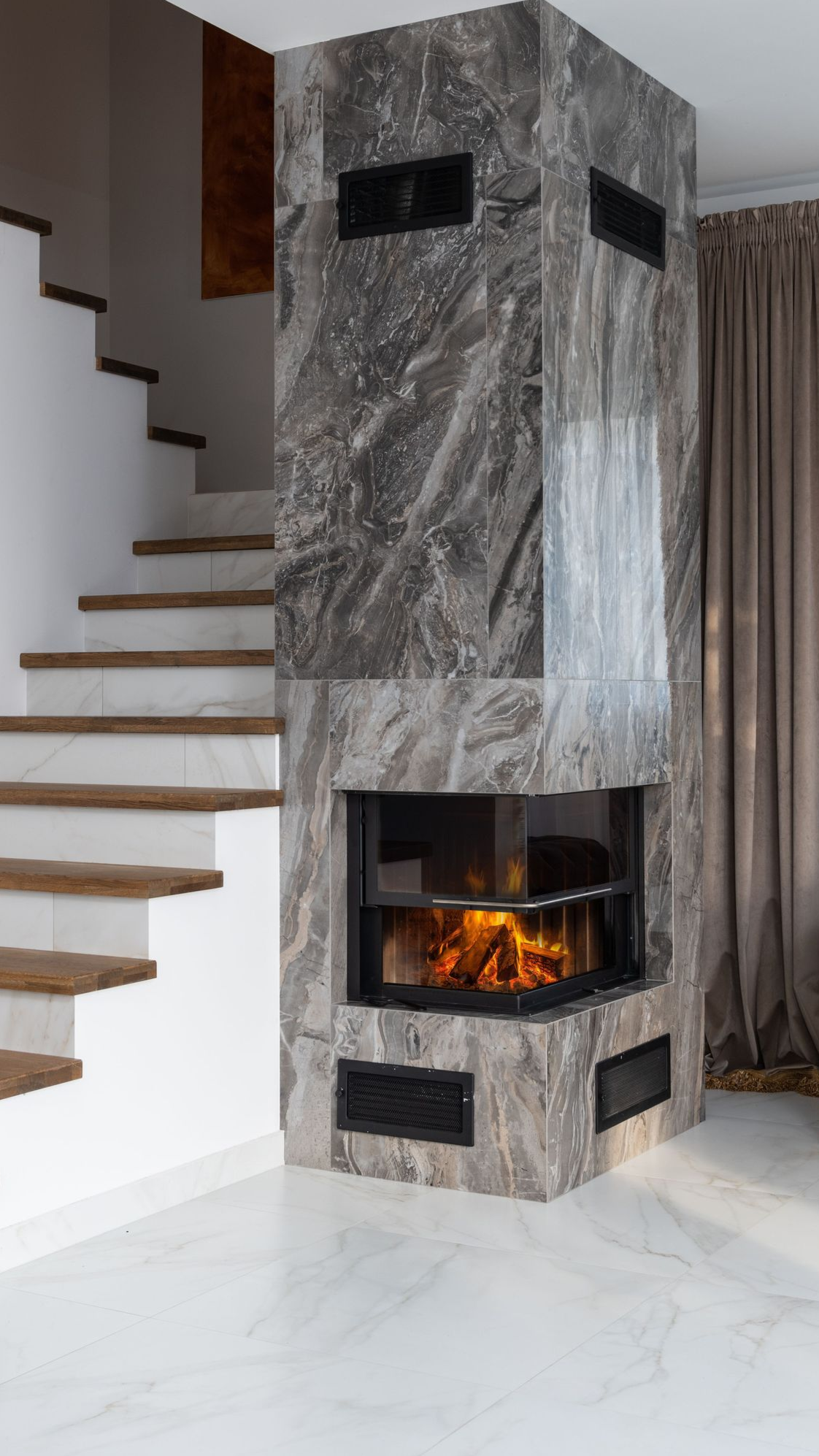 Modern corner fireplace with gray stone surround beside a white staircase and beige curtains