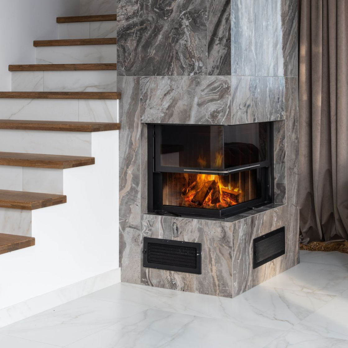 Modern corner fireplace with gray stone surround beside a white staircase and beige curtains