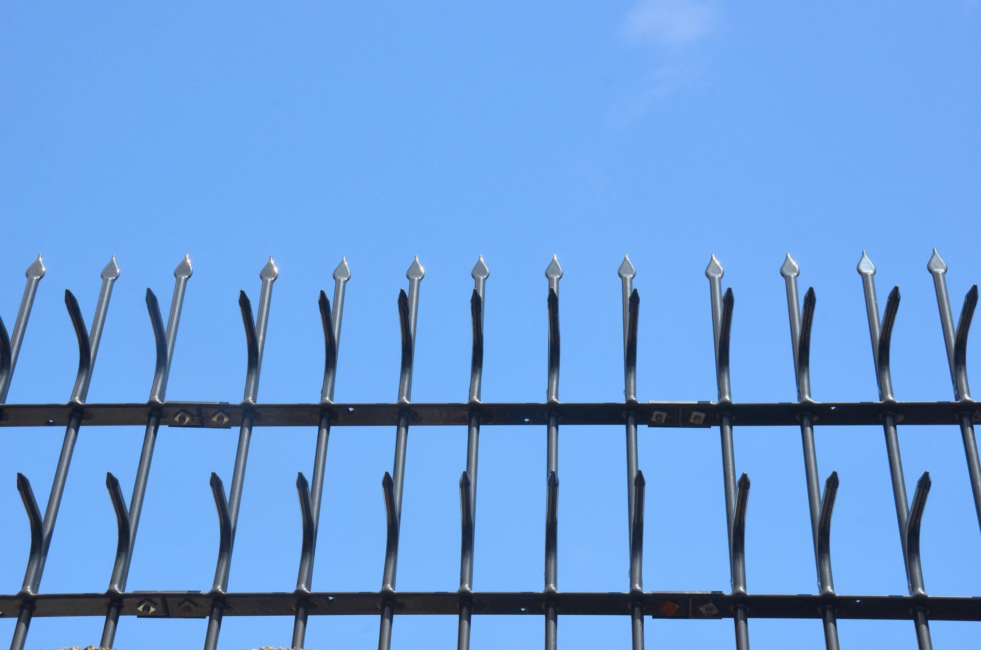 Black metal security fence with spear-top pickets silhouetted against a clear blue sky. Black metal security fence with spear-top pickets silhouetted against a clear blue sky.