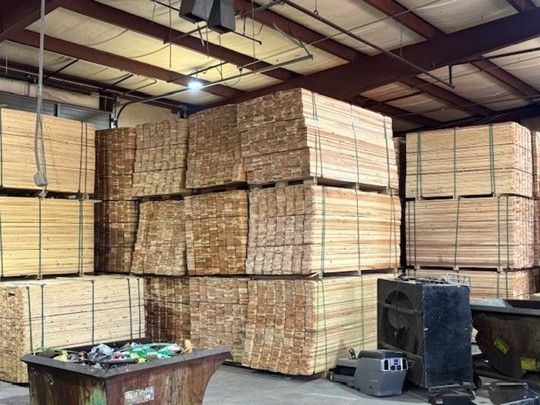 Stacks of bundled lumber inside a warehouse, with a table and equipment in the foreground.