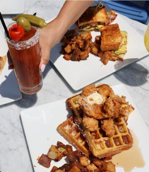 Brunch spread with chicken and waffles, sausage, and a tall red cocktail on a white table near Prelude at Medley.