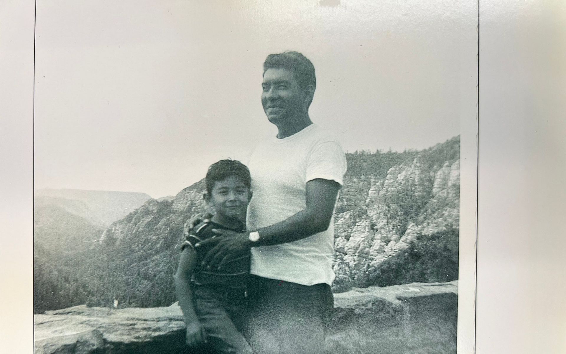 A man and a boy pose together outdoors, embracing. They stand on a rocky outcrop overlooking a vast, wooded canyon.