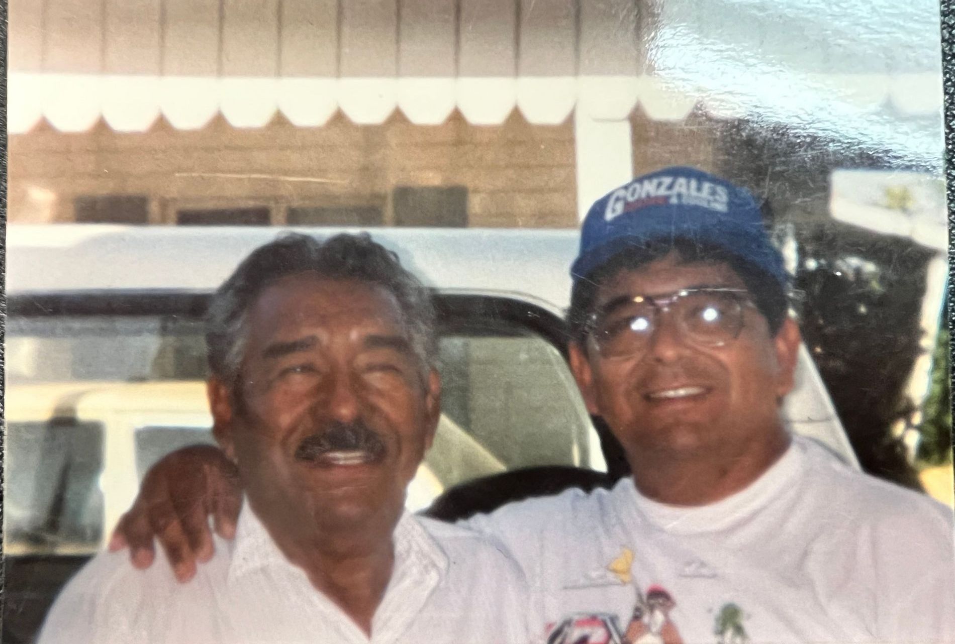 Two men smiling, one with a mustache, the other in a blue hat. They are outside a building, arm in arm, with a car visible in the background.