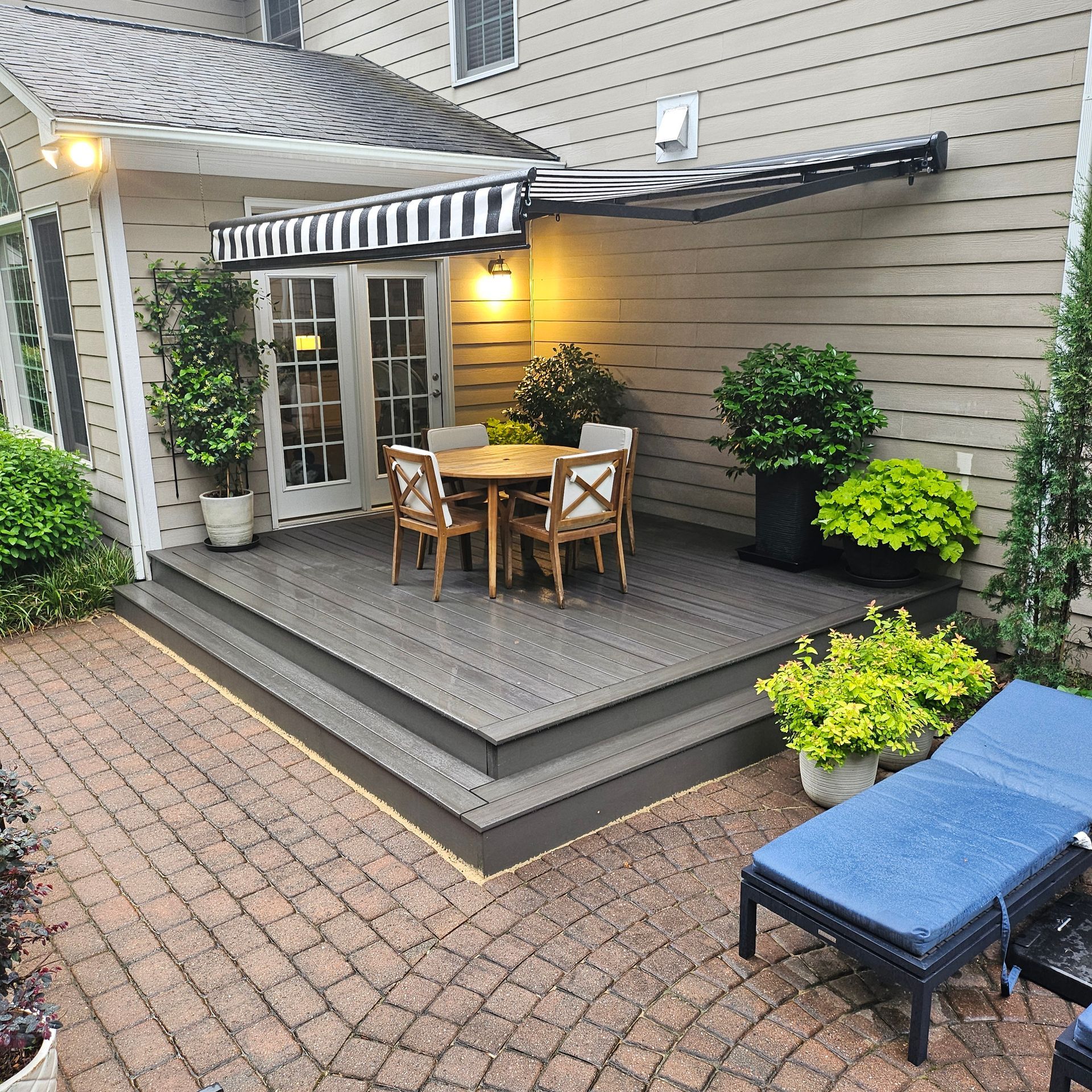 Covered backyard deck with striped awning, small dining table, potted plants, and blue lounge chair.