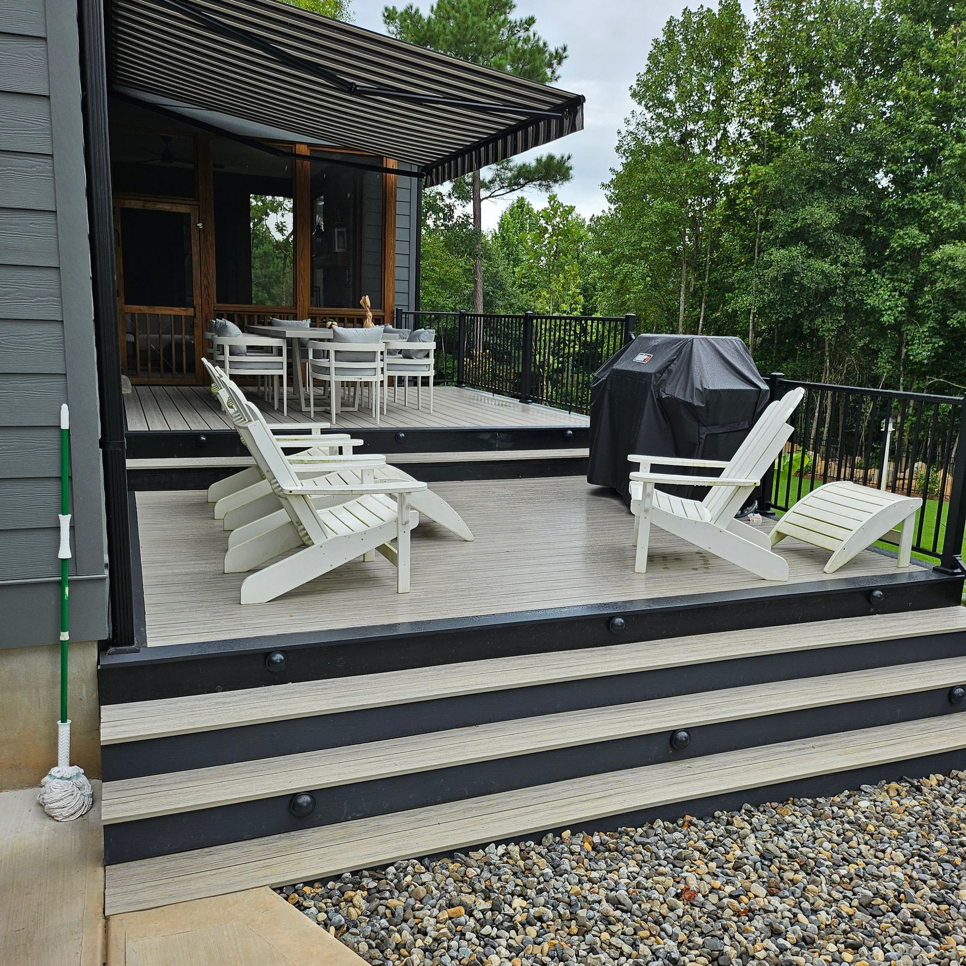 Outdoor deck with white lounge chairs, dining table, and a black grill beside a house in a green yard