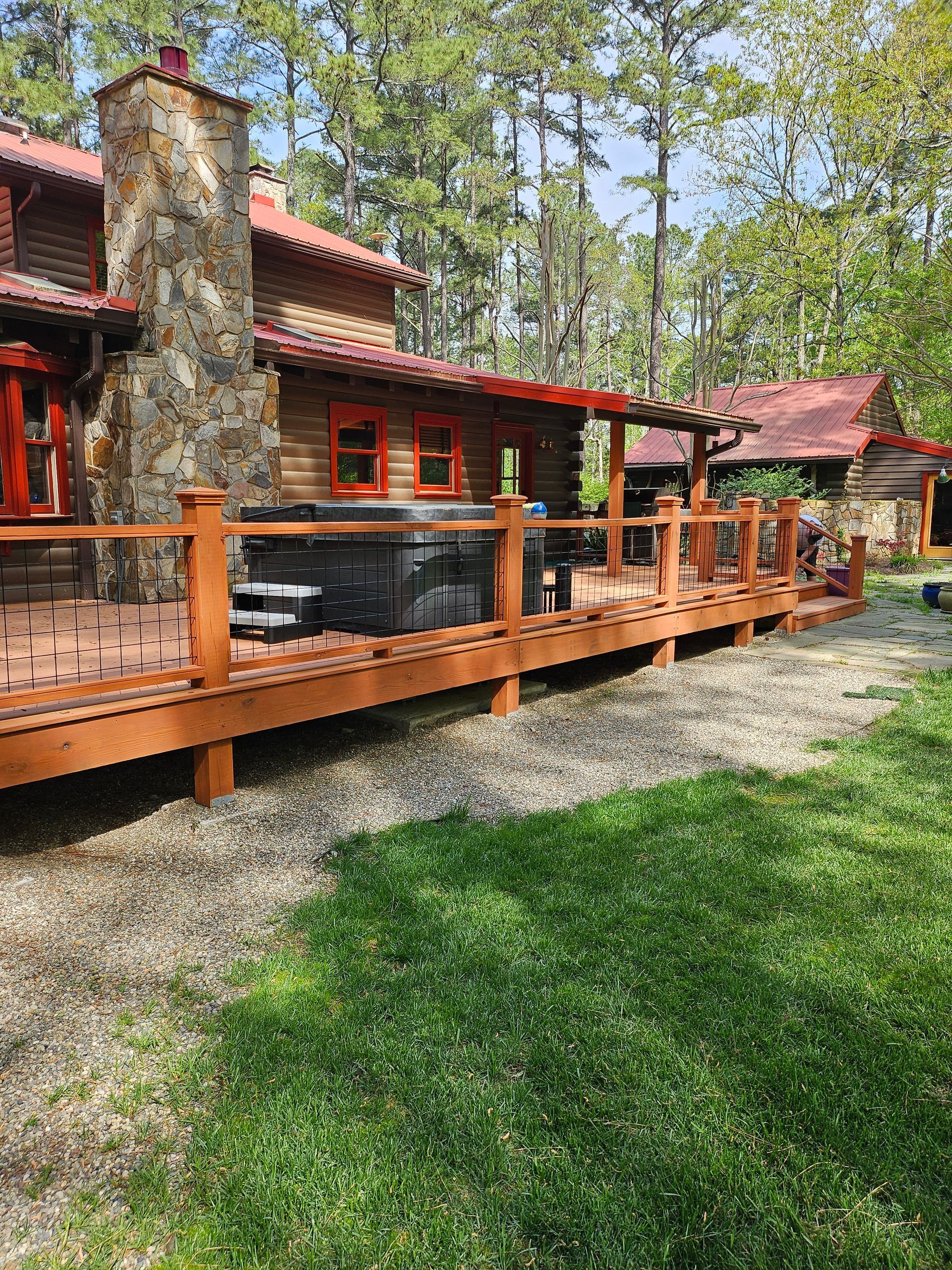 Rustic lodge with red roof, stone chimney, and wooden deck surrounded by trees and gravel path