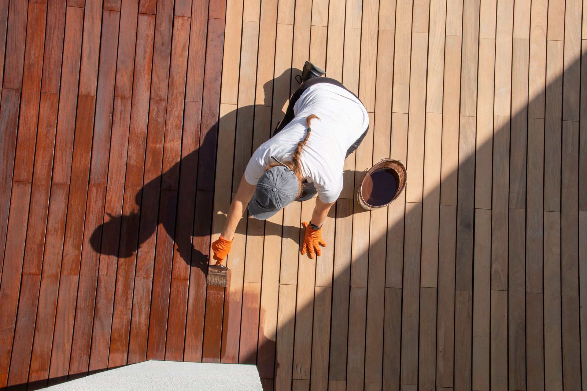 Person in white shirt and orange gloves cleaning a wooden deck in sunlight.
