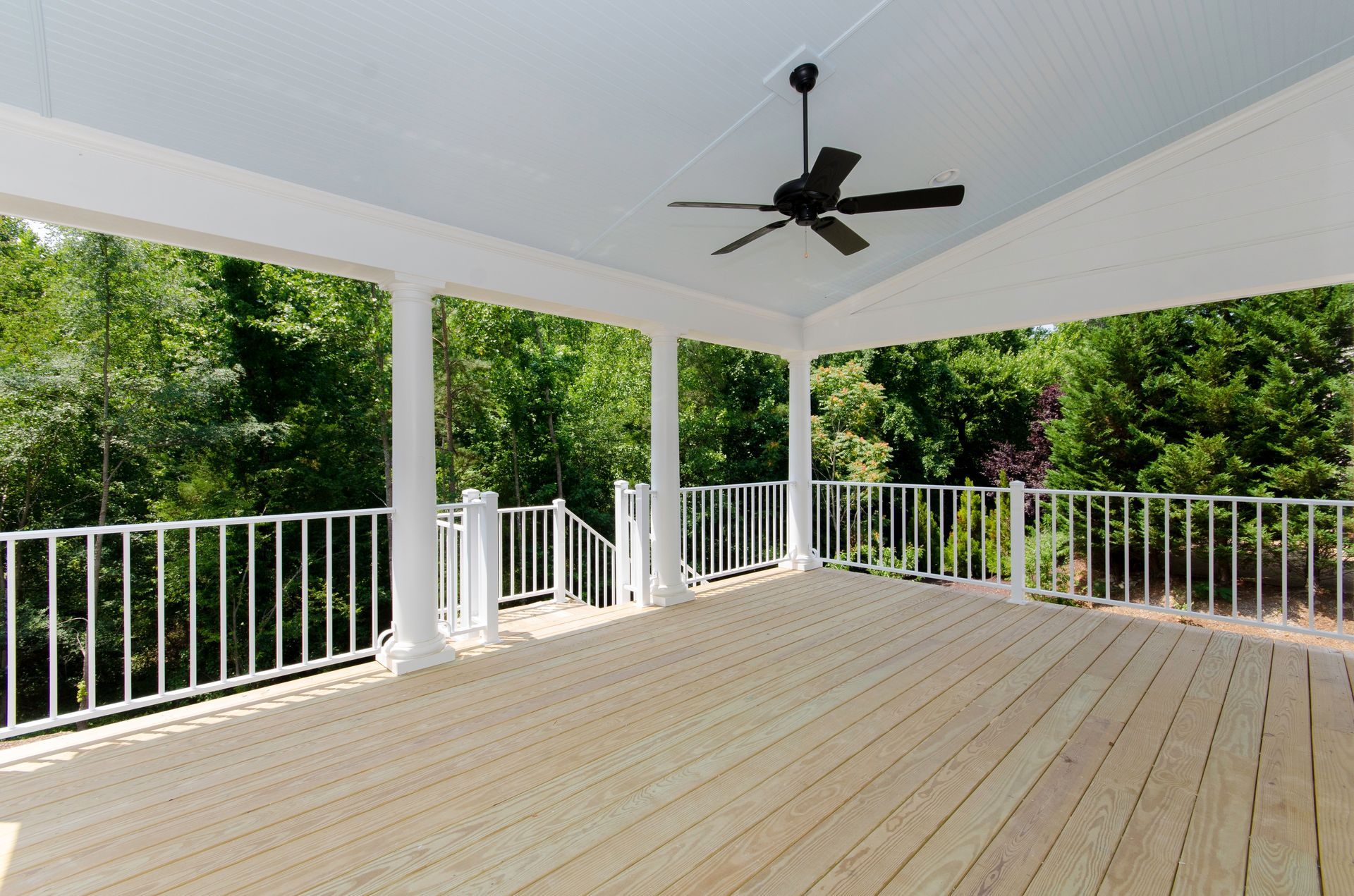 Covered wooden deck with white railing, ceiling fan, and view of green trees.