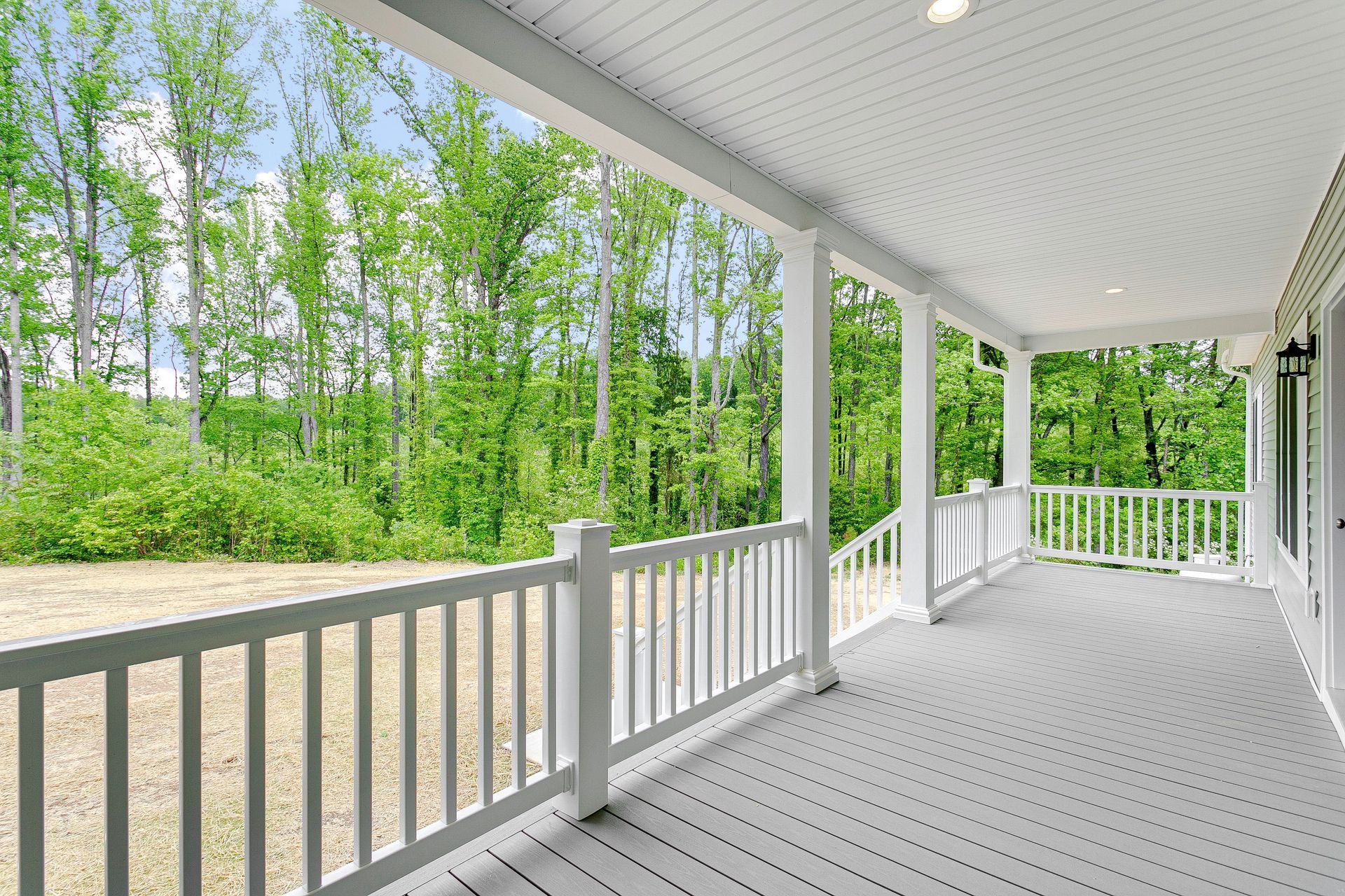 Covered porch with white railings overlooking a wooded yard and gravel driveway