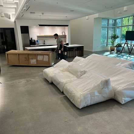 A person moving furniture in an open office space. Sofas wrapped in plastic are in the foreground.