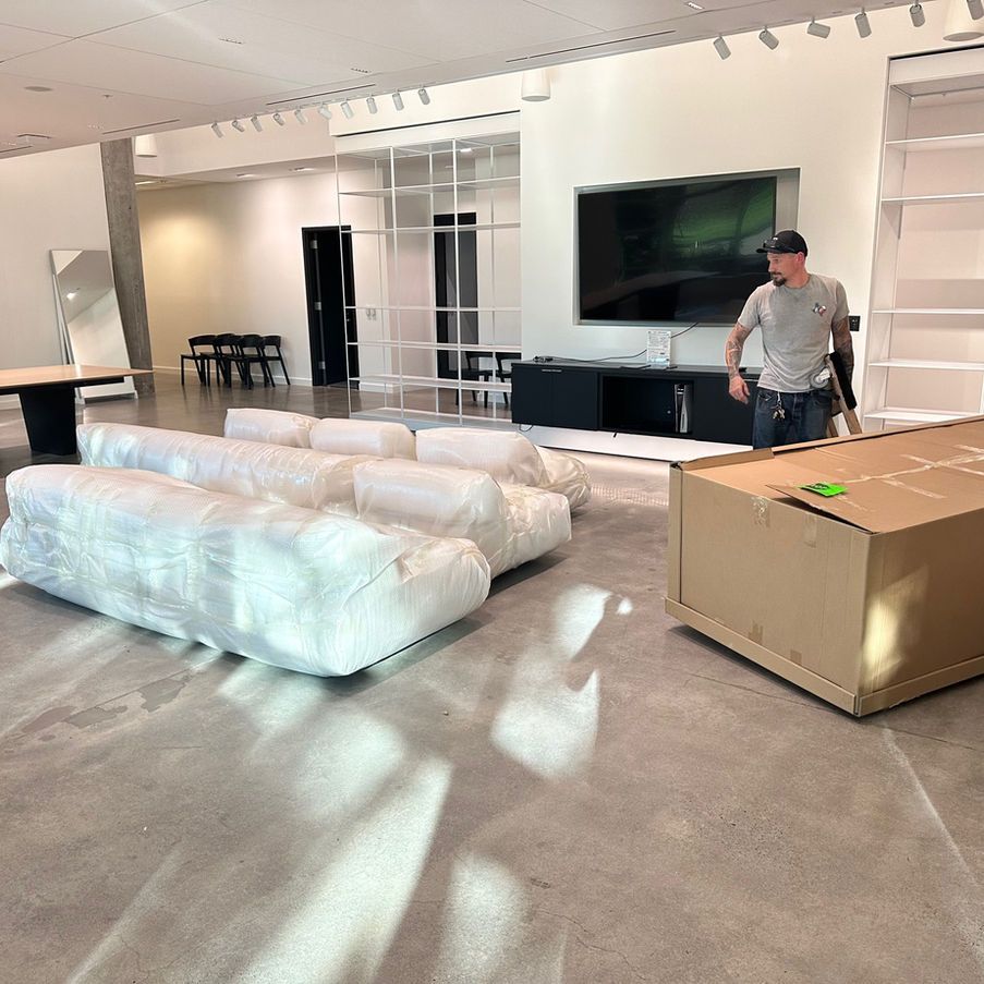 A man standing in a large room with furniture wrapped in plastic, and a cardboard box.