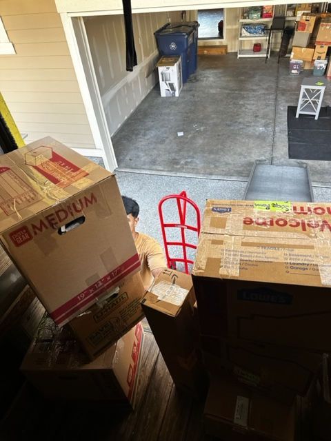 Person unloading moving boxes into a garage. Boxes are stacked near a red hand truck.