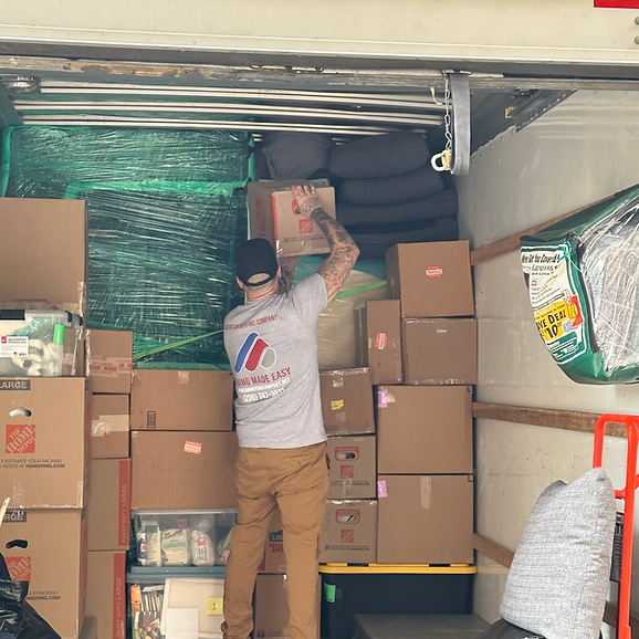 Person in a moving truck stacking boxes. Interior shot with tan boxes, green wrapping, and a gray-shirted person.