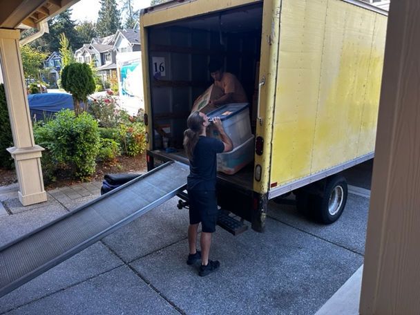 Man loading a box and items into a yellow moving truck on a driveway.