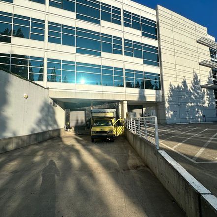 Yellow truck parked under a modern building's overhang. Shadows from trees and sun glare.