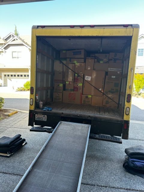 Yellow moving truck loaded with boxes, ramp extended. Parked in a driveway.