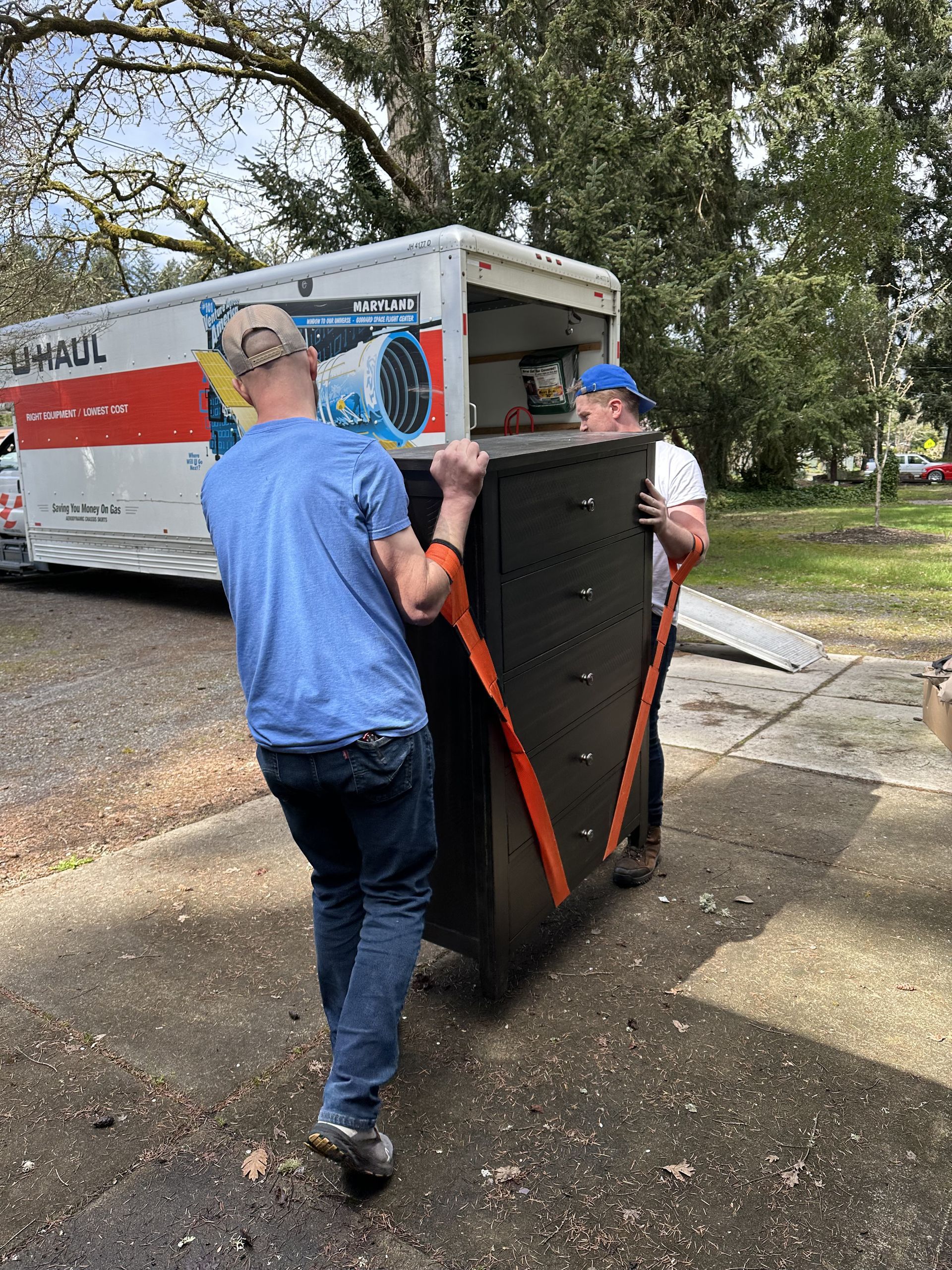 Two people carrying a dark dresser with orange straps towards a U-Haul truck on a driveway.