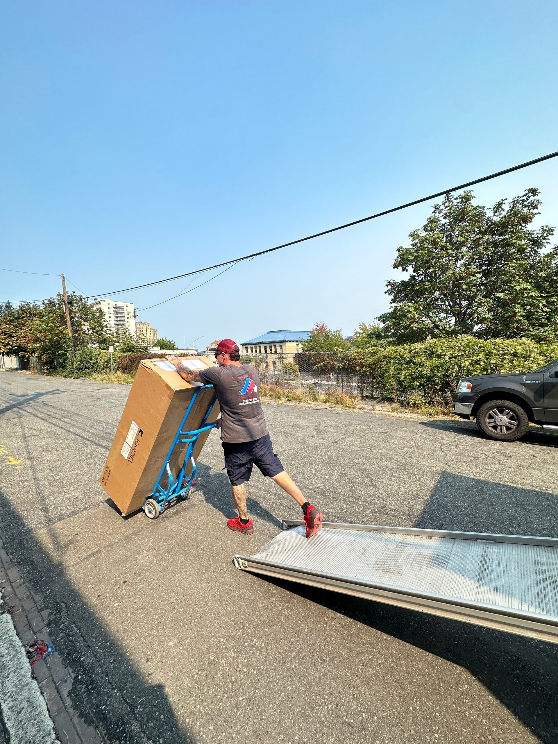 Man pushing a large cardboard box on a dolly up a ramp on a gravel driveway.