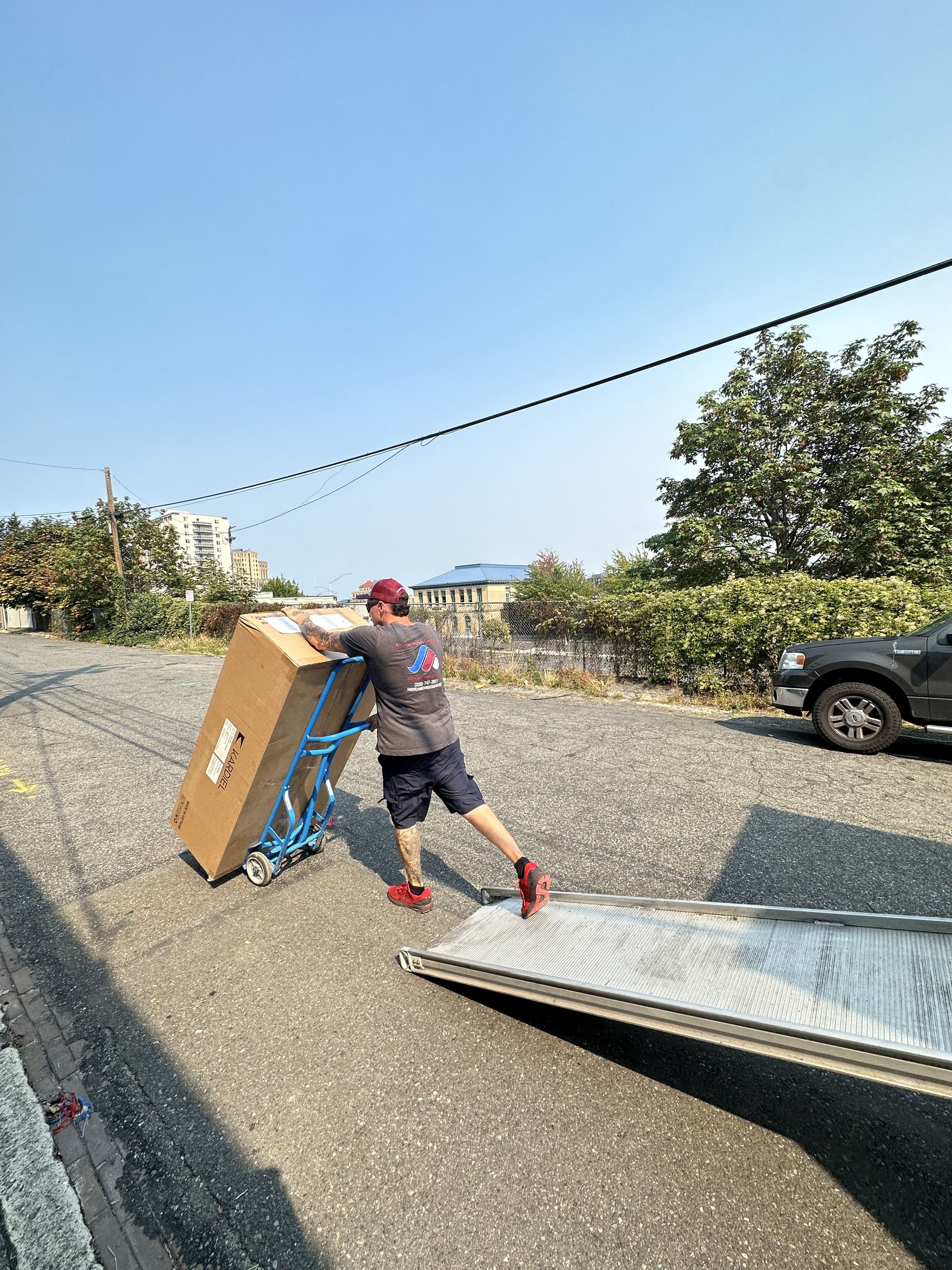 Person pushing a large box on a dolly toward a ramp on a gravel surface. Sunny outdoor setting.