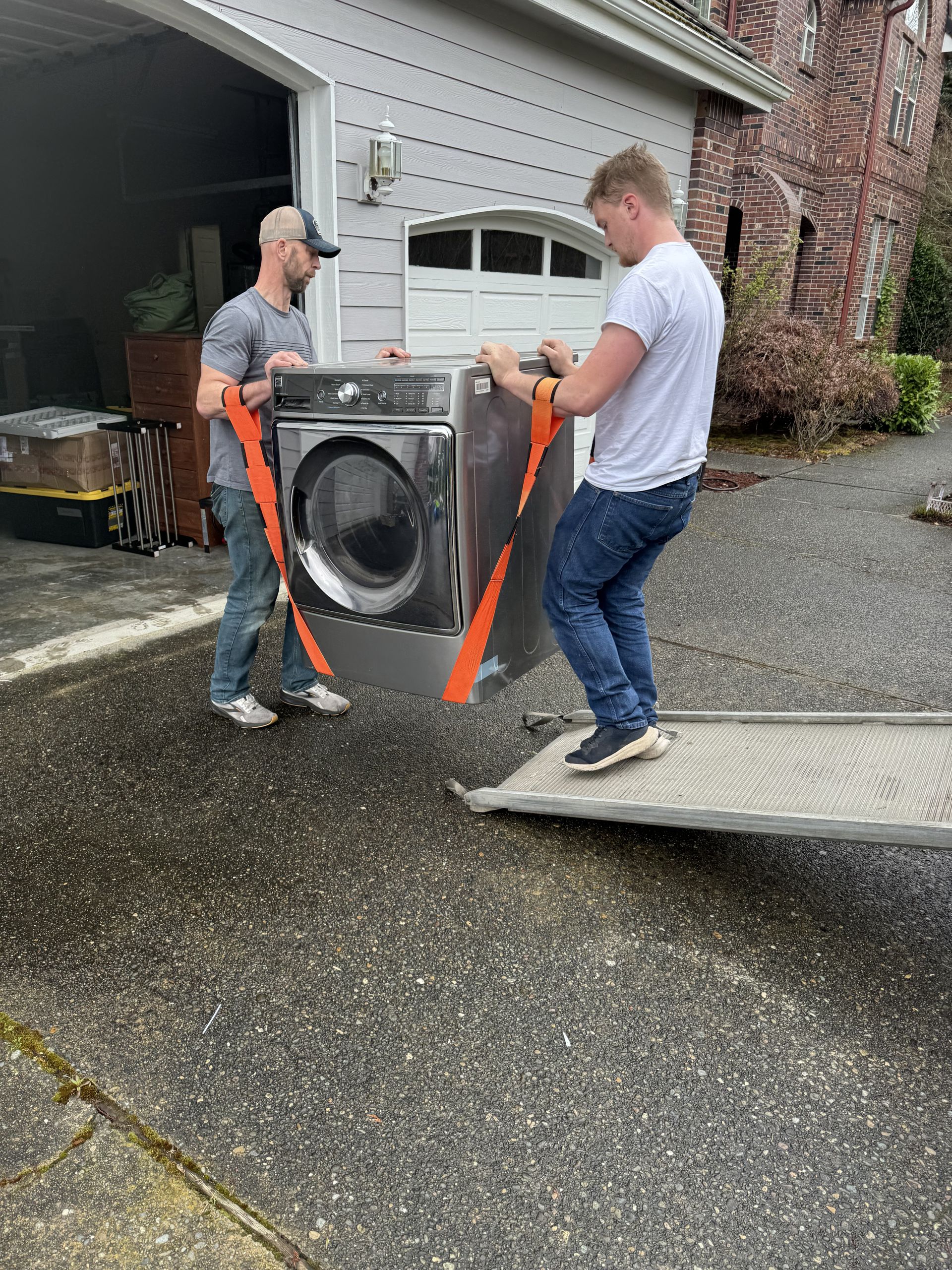 Two people use straps to load a washing machine onto a ramp, near a garage.