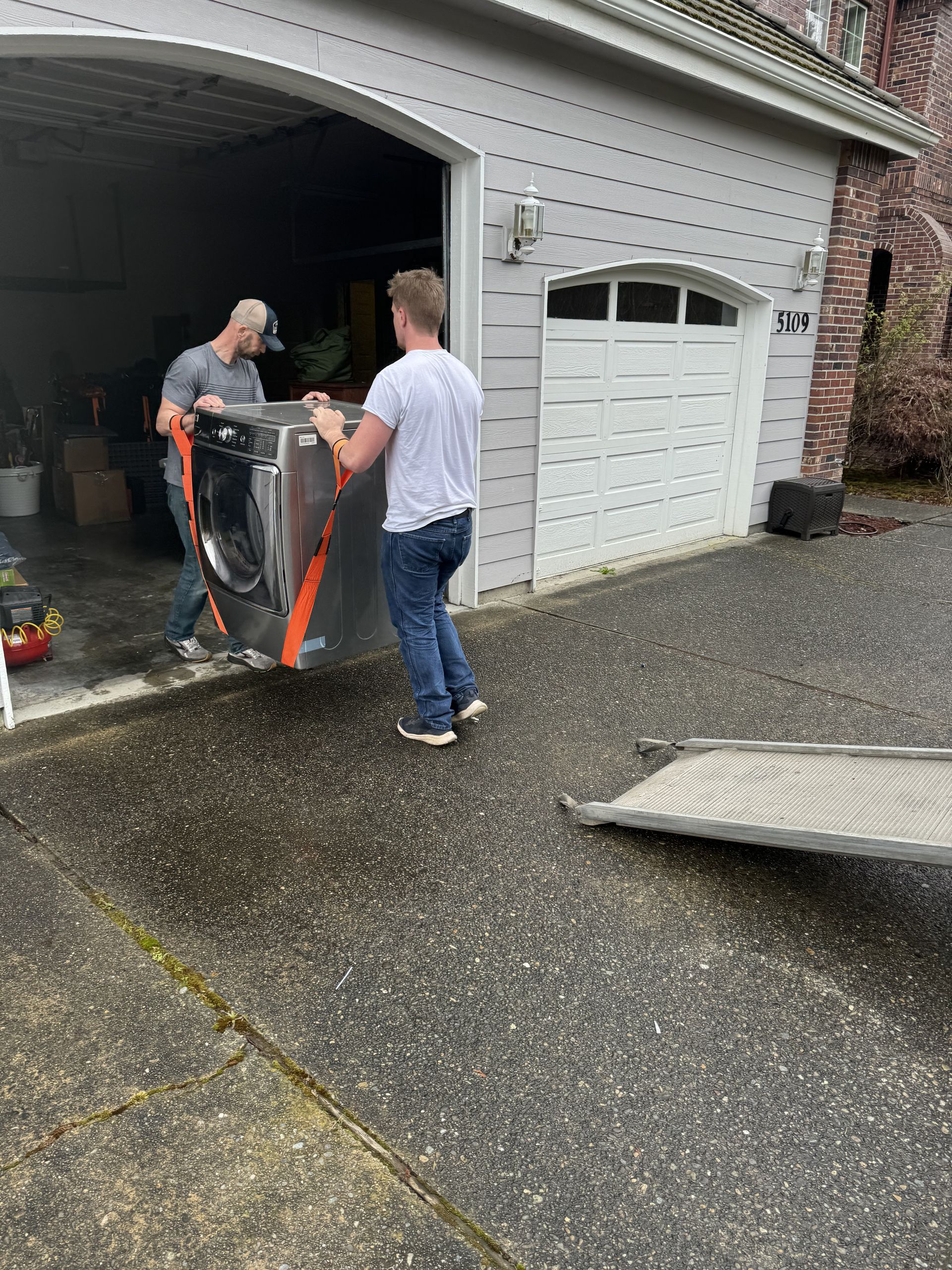 Two people carrying a large, wrapped item out of a garage. The item is silver and black.
