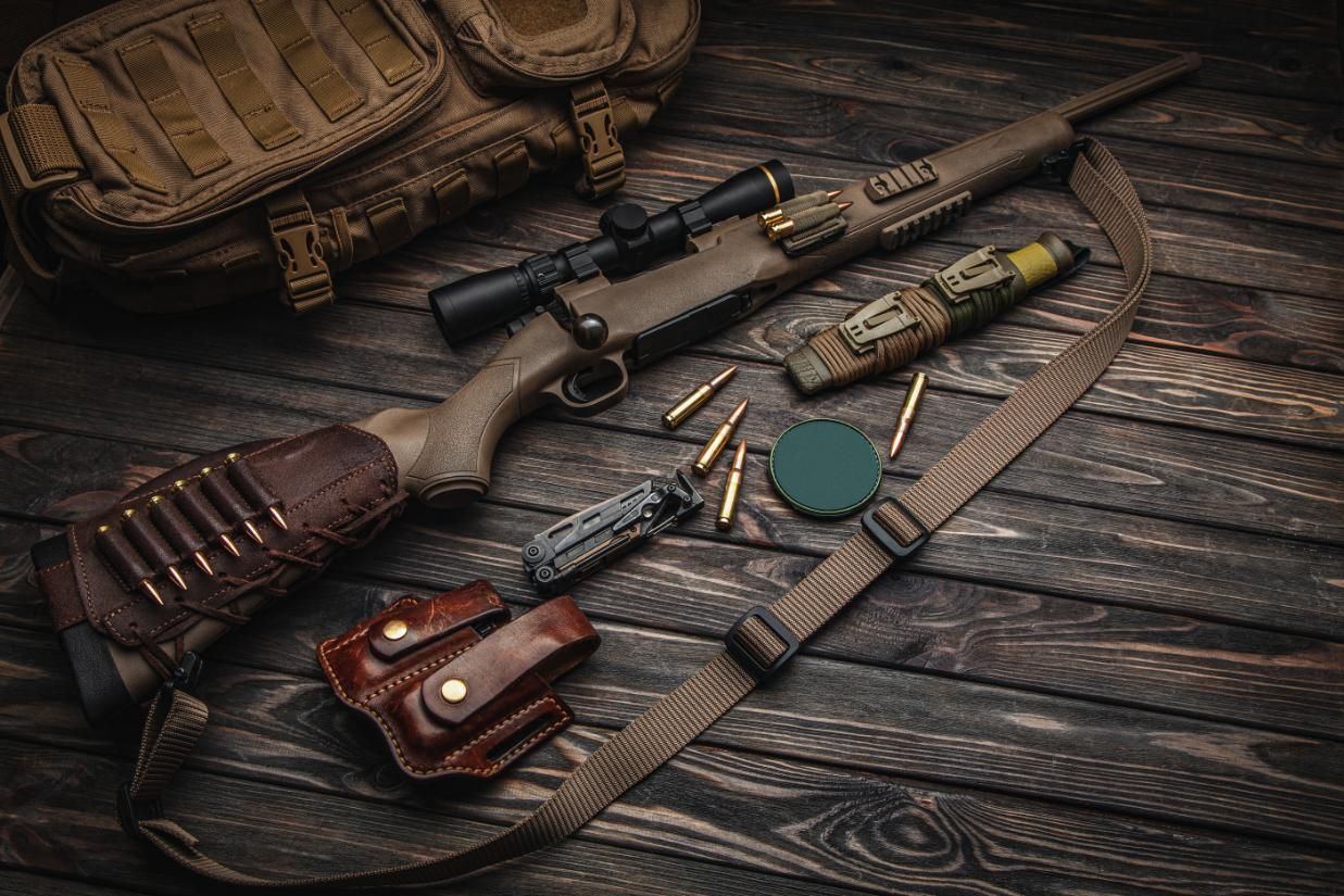 Tactical rifle with scope, ammo, knife, holster, sling, and bag on a wooden surface.